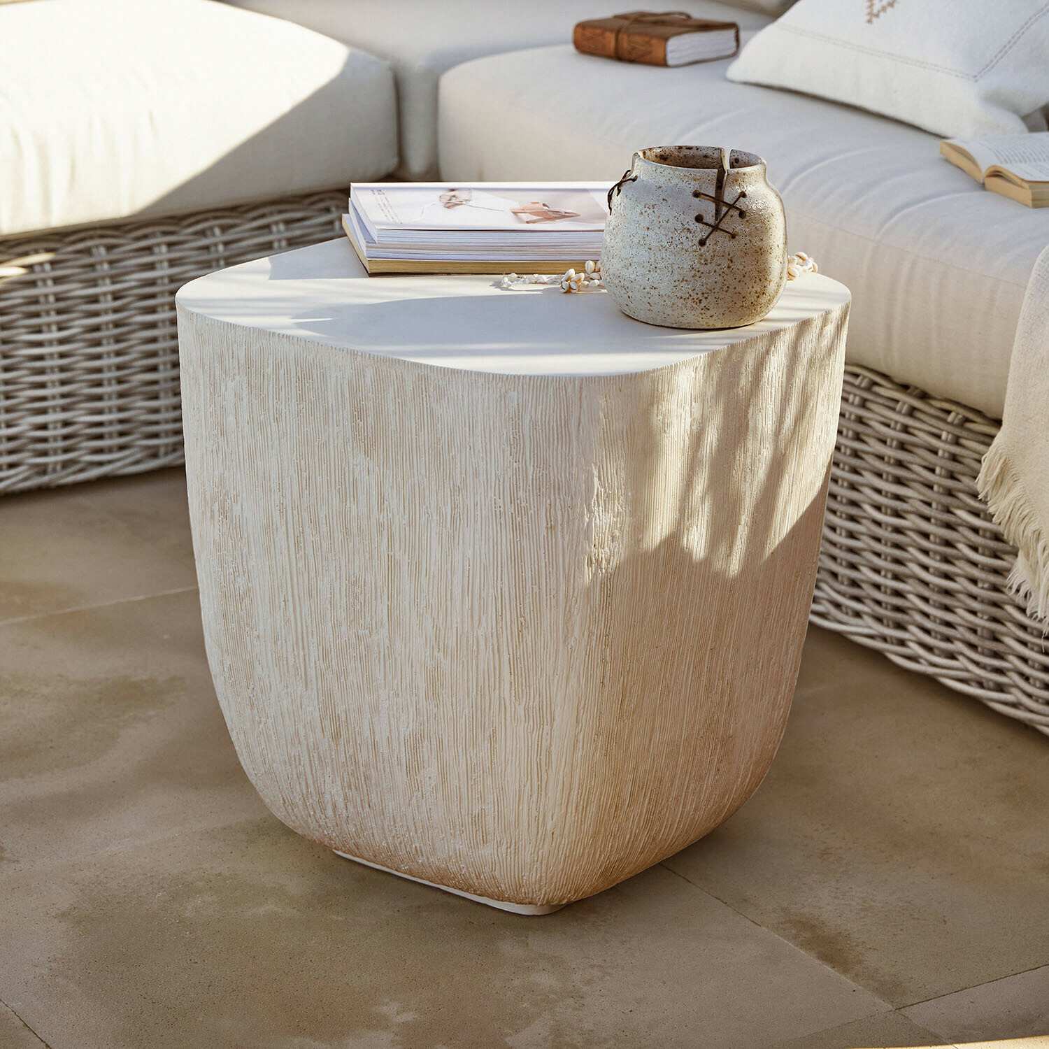 Table d'appoint beige texturée effet bois avec pot en céramique et livres sur terrasse avec canapé en osier