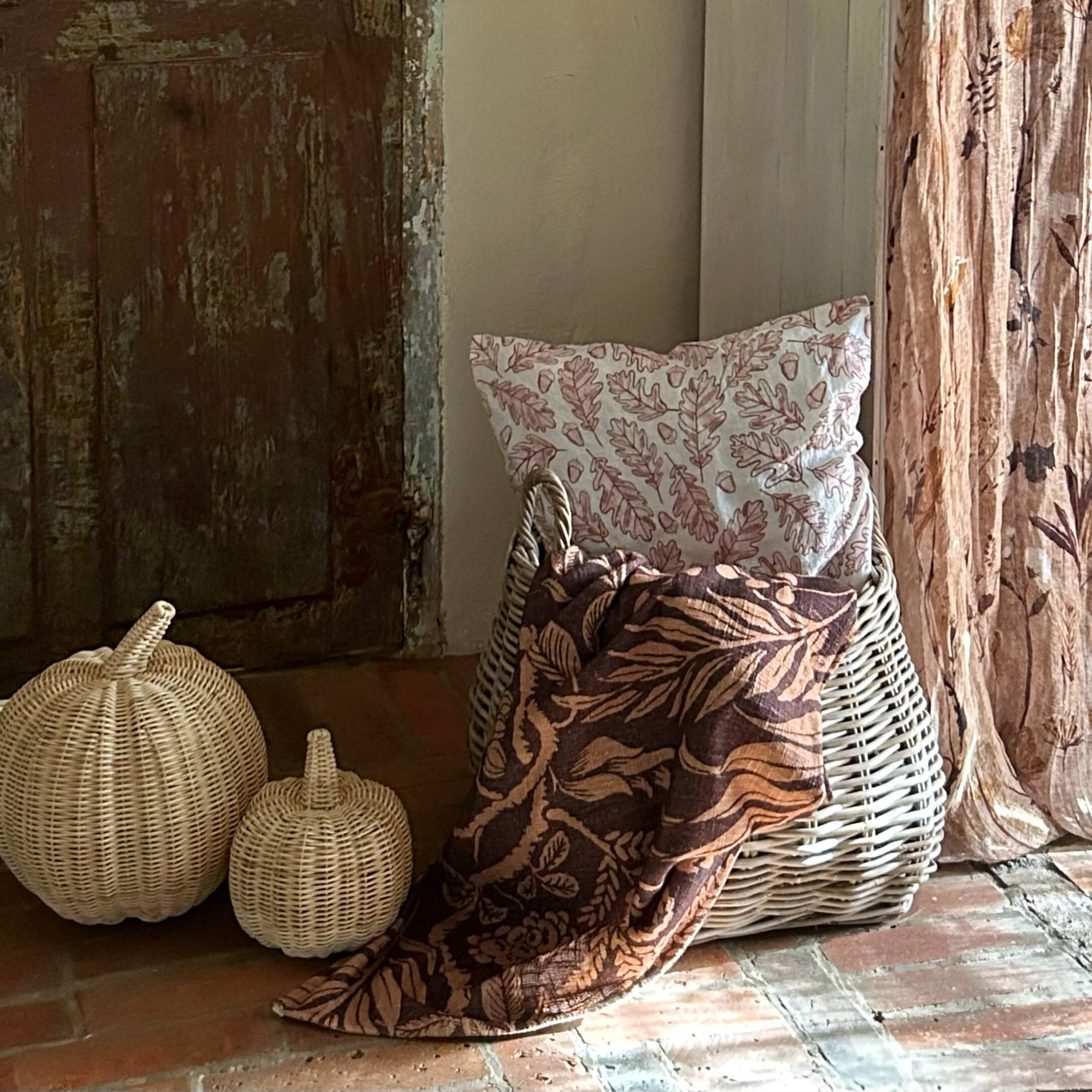 Panier en osier avec coussin à motif feuilles et couverture marron à motifs floraux sur sol en briques