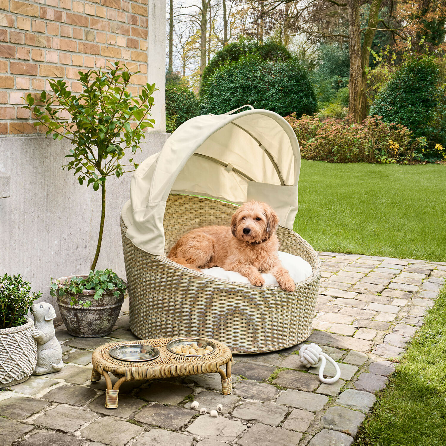 Panier rond en rotin beige avec auvent crème et coussin pour chien dans un jardin pavé