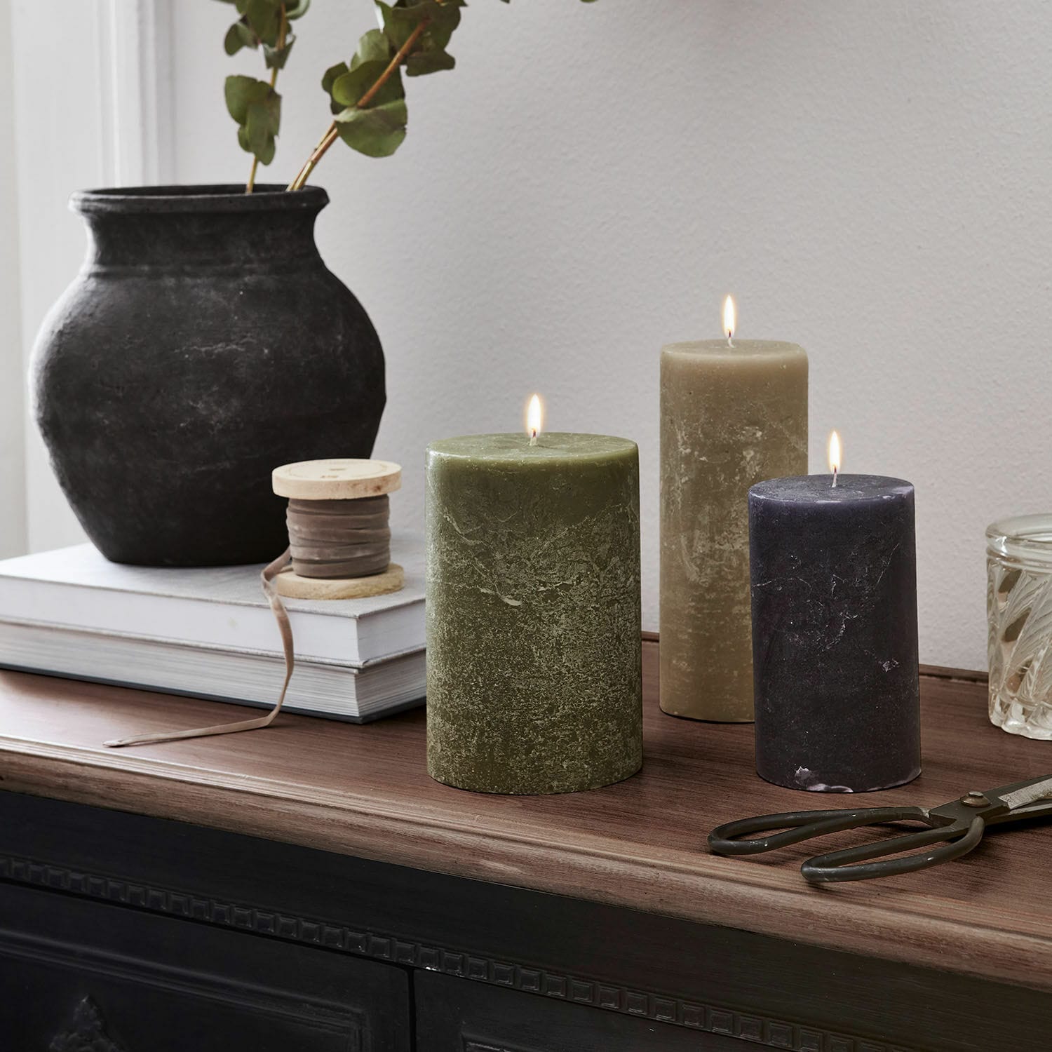 Lot de 3 bougies Pretin Trois bougies allumées vert, beige et noir sur un plateau en bois avec décorations d'intérieur