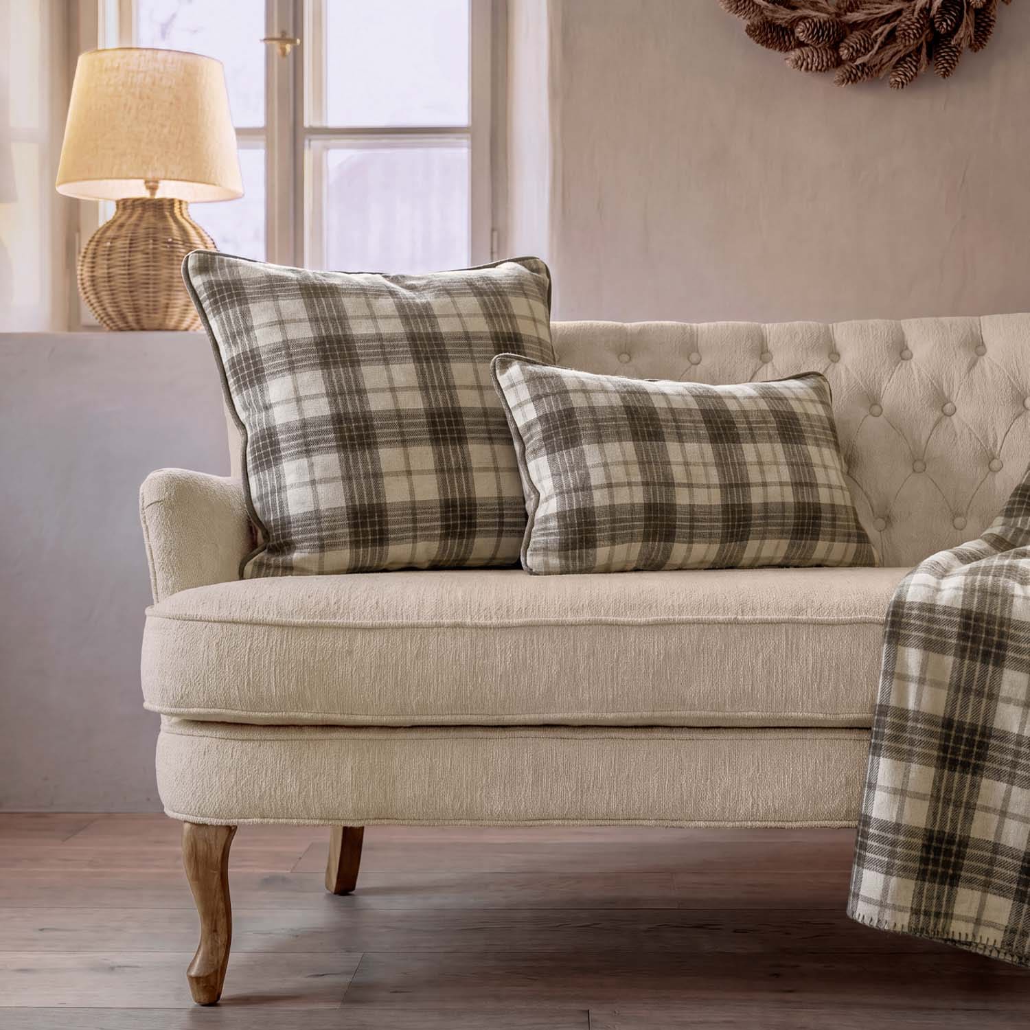 Canapé beige avec deux coussins à carreaux gris et crème, lampe en osier près d'une fenêtre