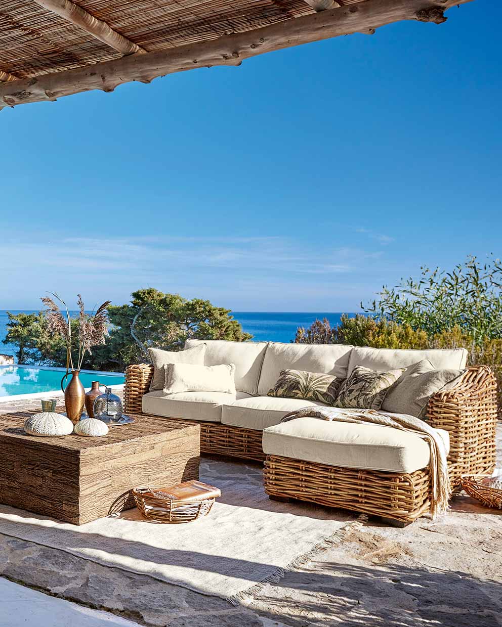 Ensemble de mobilier d'extérieur en rotin avec coussins crème et table en bois sur terrasse avec vue sur la mer