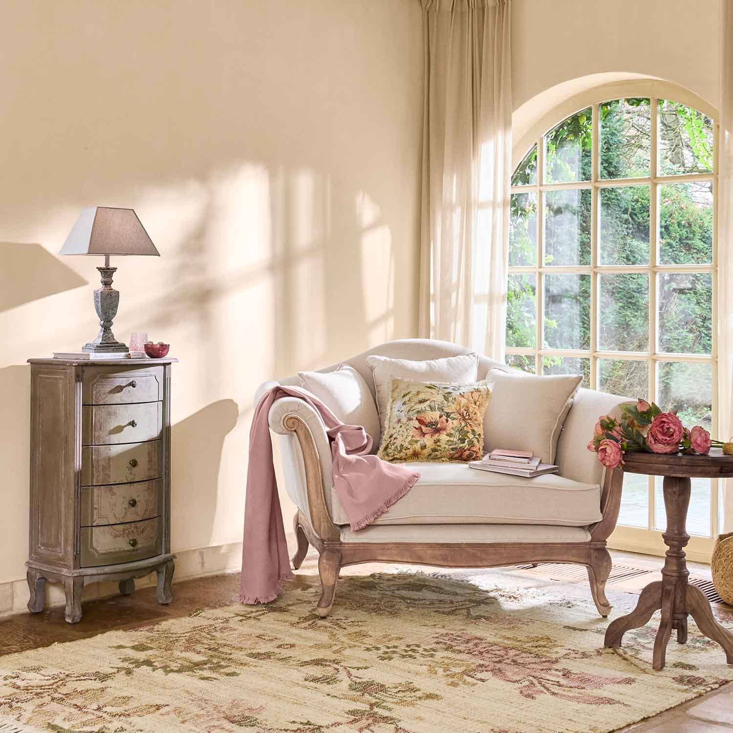 Canapé crème avec cadre en bois, coussin floral et plaid rose dans un salon lumineux avec grande fenêtre