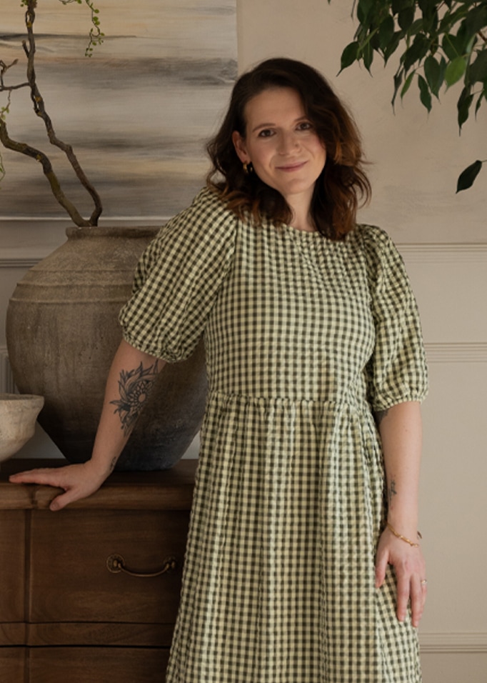 Femme en robe à carreaux verts appuyée sur une commode en bois avec un grand vase, intérieur chaleureux