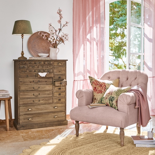 Fauteuil rose douillet avec coussins floraux à côté d'une commode en bois dans un salon lumineux aux rideaux roses
