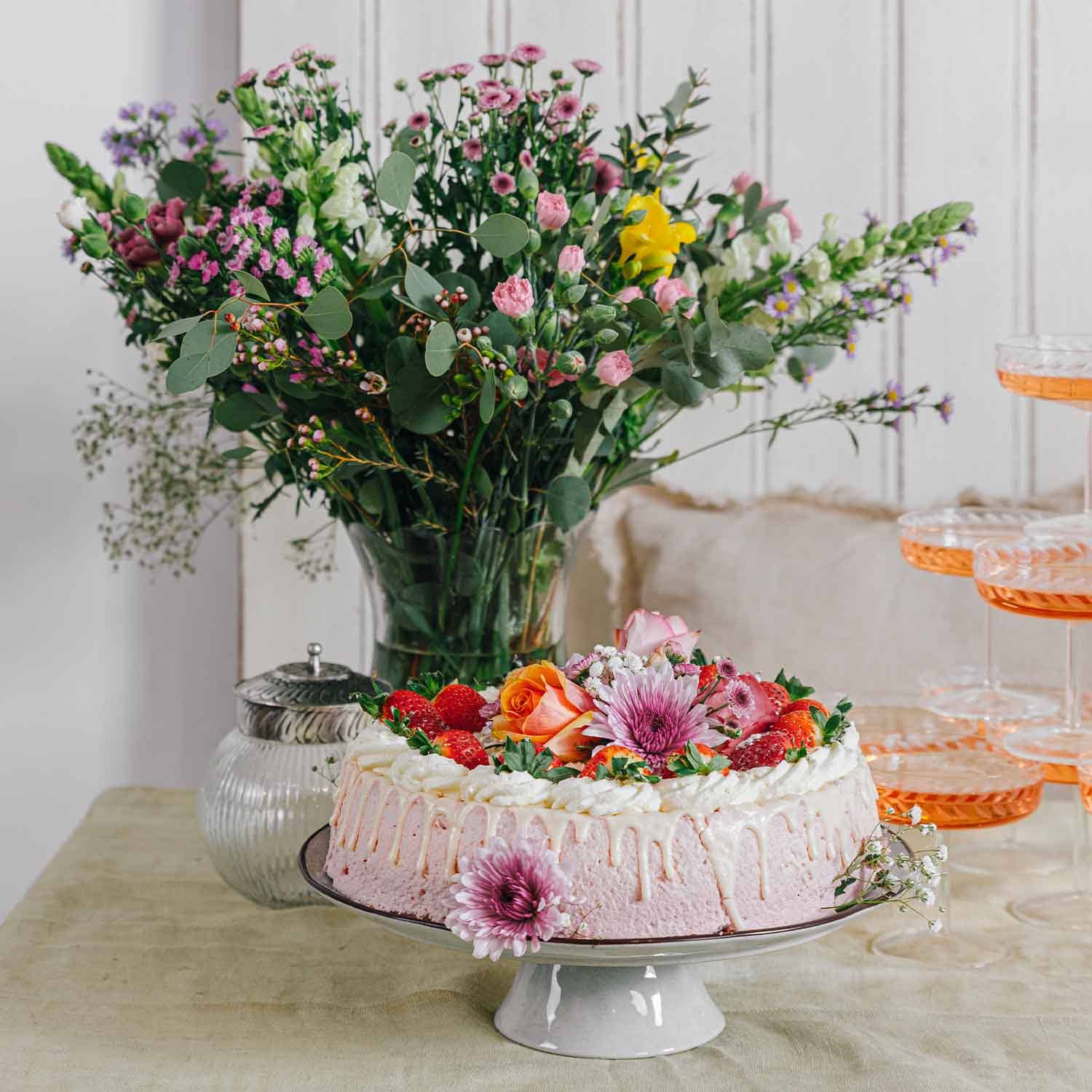 Gâteau à la crème rose décoré de fraises et de fleurs fraîches sur un présentoir blanc avec bouquet en arrière-plan