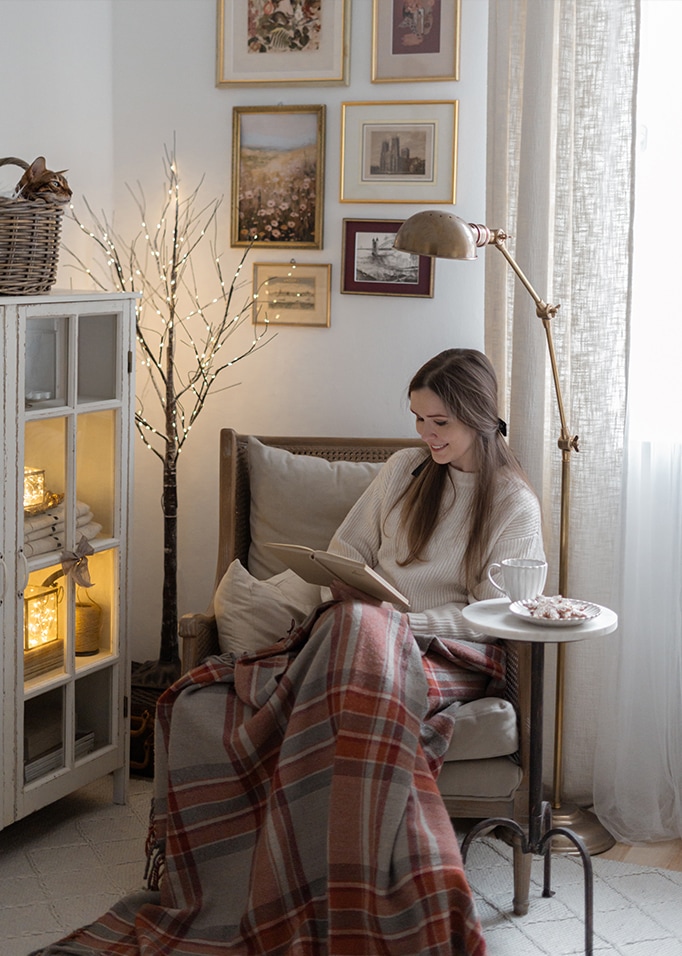 Femme lisant un livre sous une couverture chaude dans un salon cosy avec lampe et décorations murales