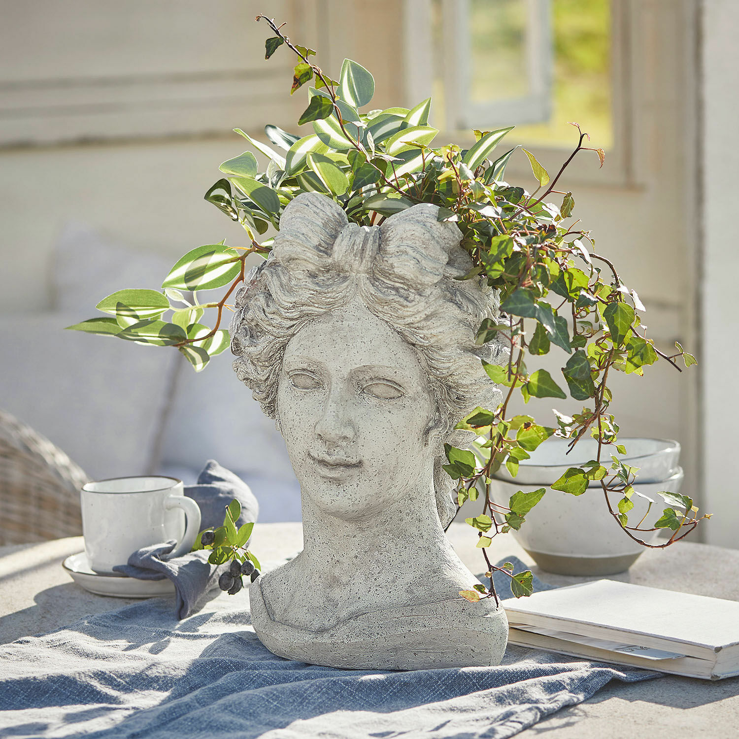Cache-pot Cassiopeia Cache-pot en forme de tête de femme en pierre grise avec plantes vertes sur une table dans une pièce lumineuse