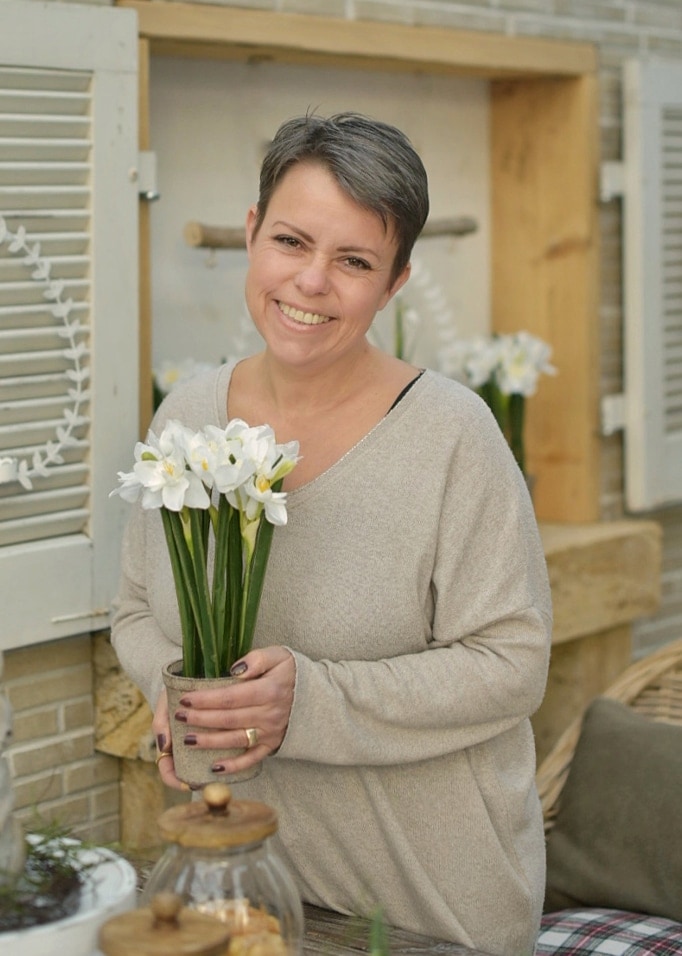 Femme en pull beige tenant un pot de fleurs blanches dans une ambiance naturelle de Pâques
