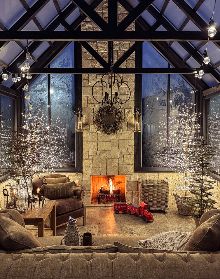 Salon chaleureux avec cheminée, mur en pierre et décorations de Noël dans une orangerie aux grandes fenêtres