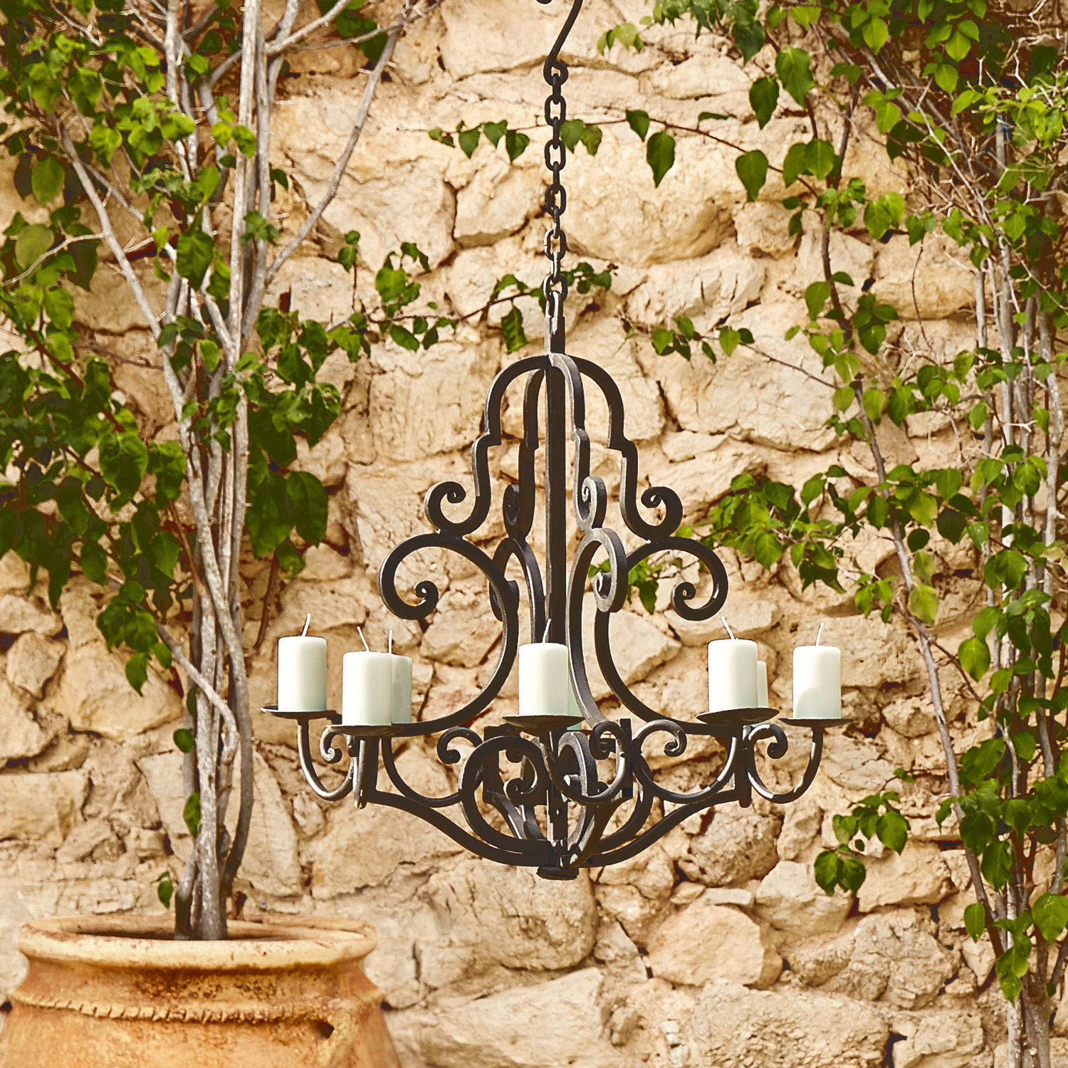 Chandelier Geneuve Lustre en métal noir avec six bougies blanches suspendu devant un mur en pierre et des feuilles vertes