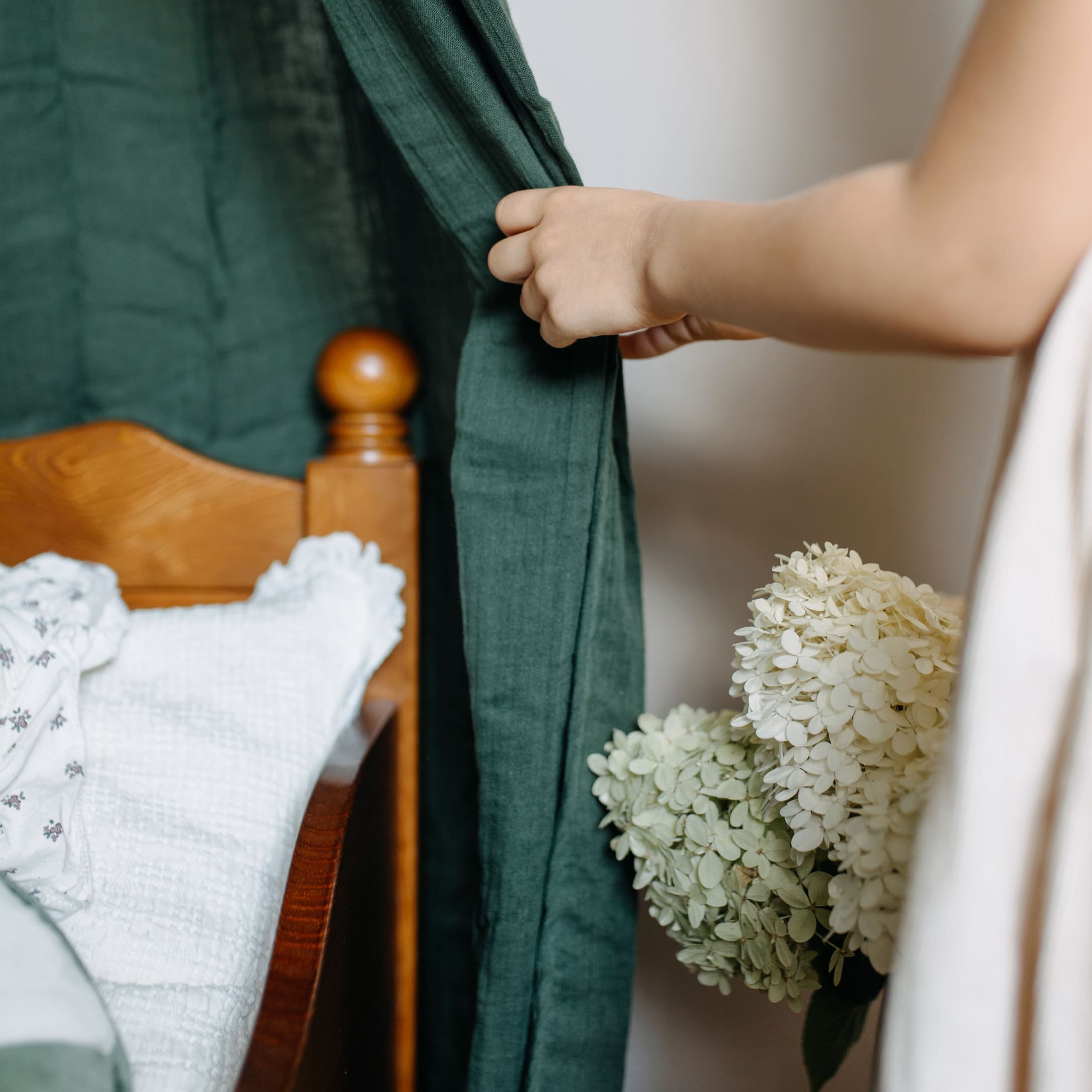 Enfant tenant un rideau vert près d'un lit en bois avec une literie blanche et un bouquet de fleurs blanches