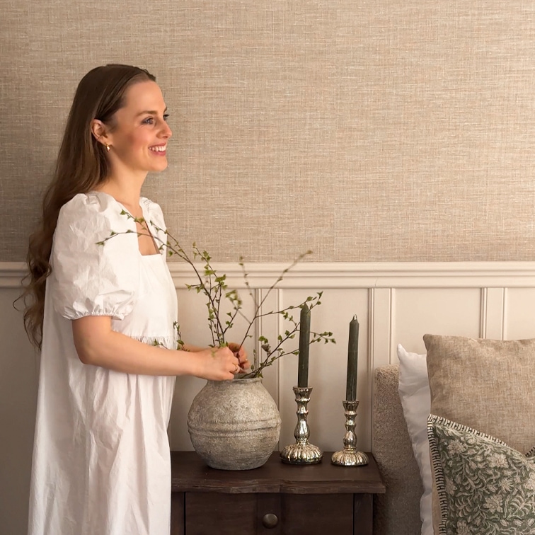 Femme en robe blanche arrange des branches dans un vase en céramique grise sur une table en bois avec des bougies