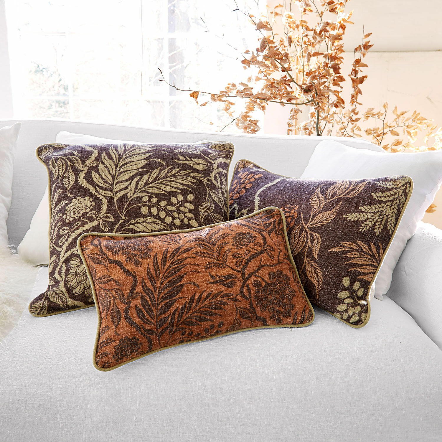 Lot de 3 housses de coussins Carivelle Ensemble de trois coussins décoratifs avec motif de feuilles en tons marron et orange sur un canapé blanc