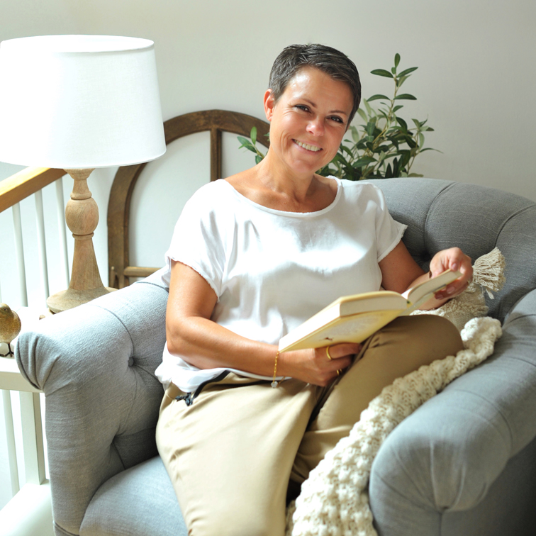 Femme en haut blanc et pantalon beige lisant un livre sur un canapé gris confortable dans une pièce lumineuse
