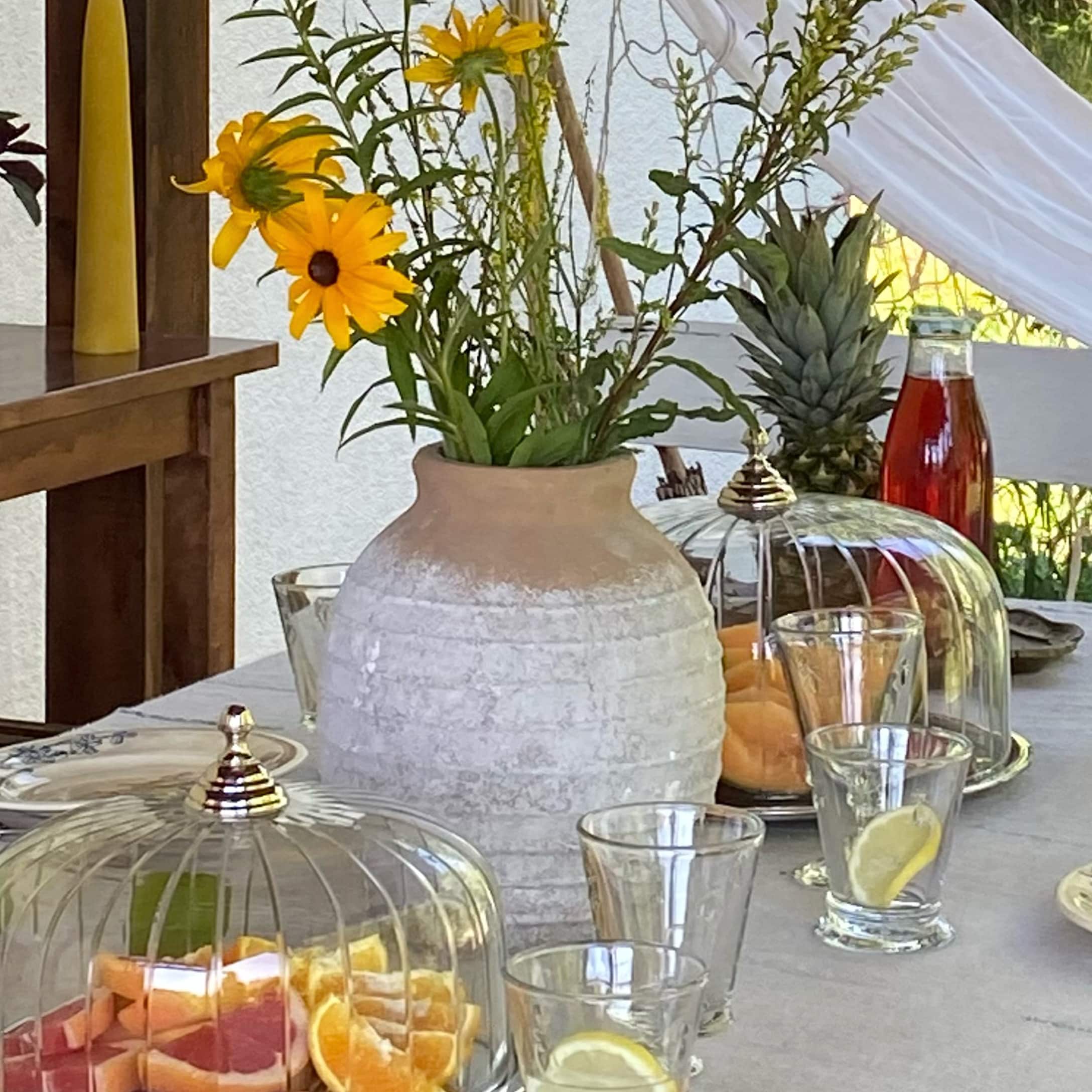 Vase en céramique beige et blanc avec fleurs jaunes sur table avec fruits et verres.
