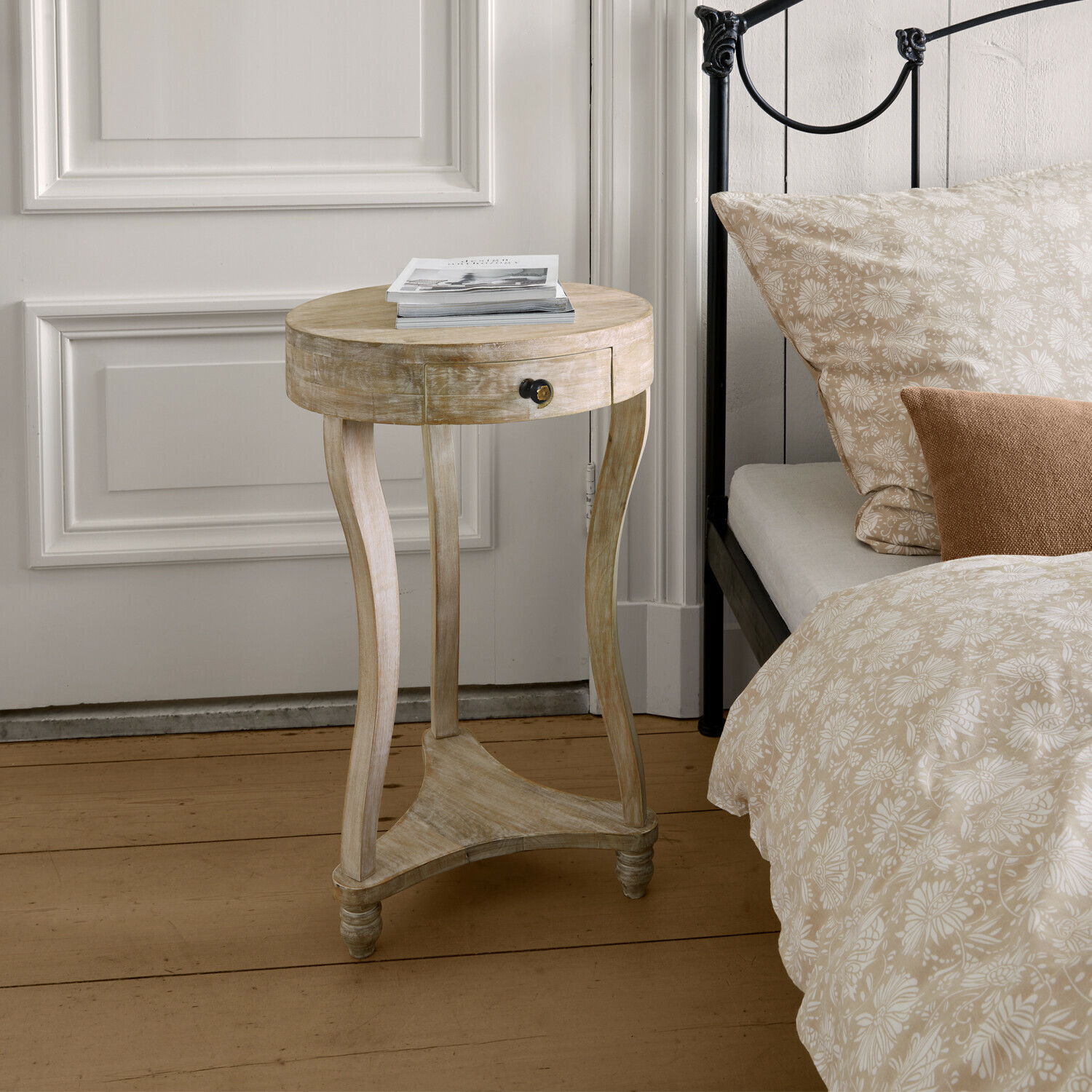 Table d’appoint Elmbury Table de chevet ronde en bois clair avec tiroir près d'un lit avec literie beige à motifs floraux