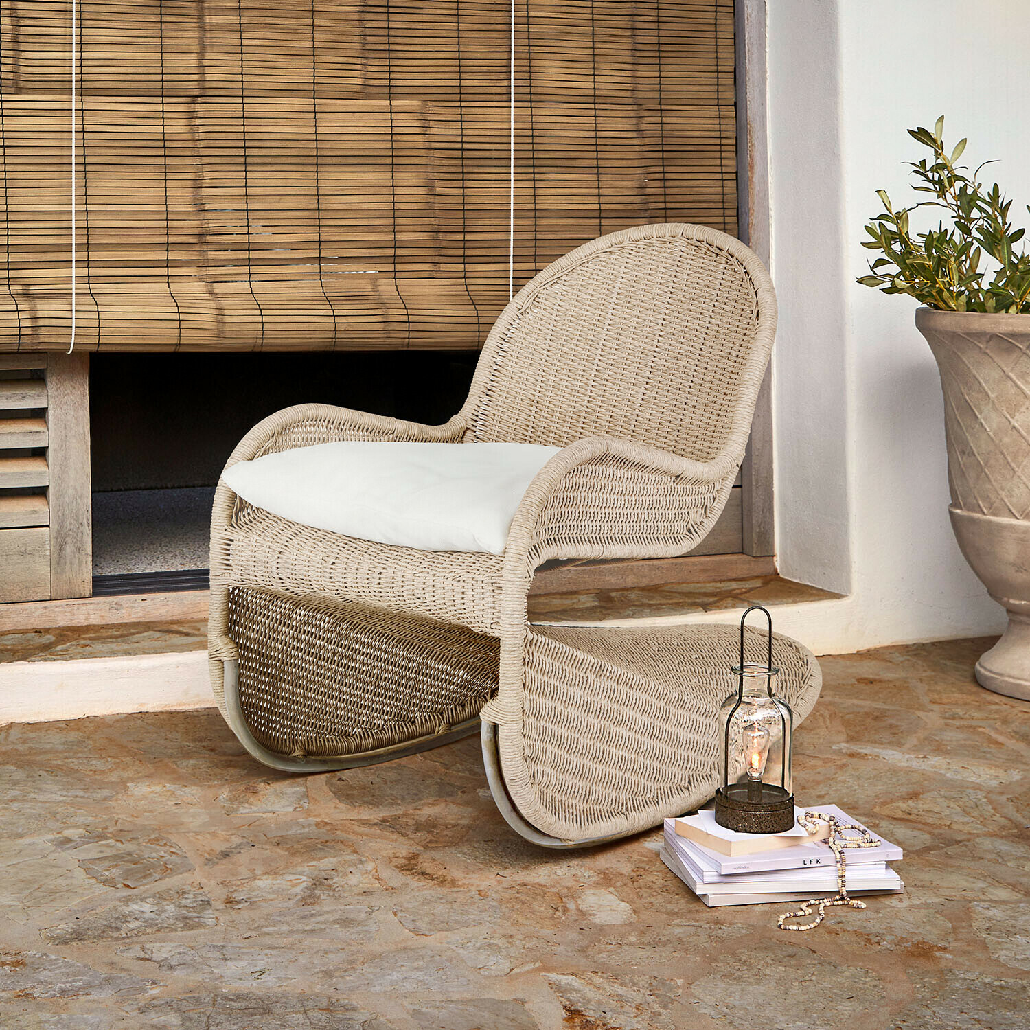 Fauteuil en rotin beige avec coussin blanc sur sol en pierre devant un store en bambou
