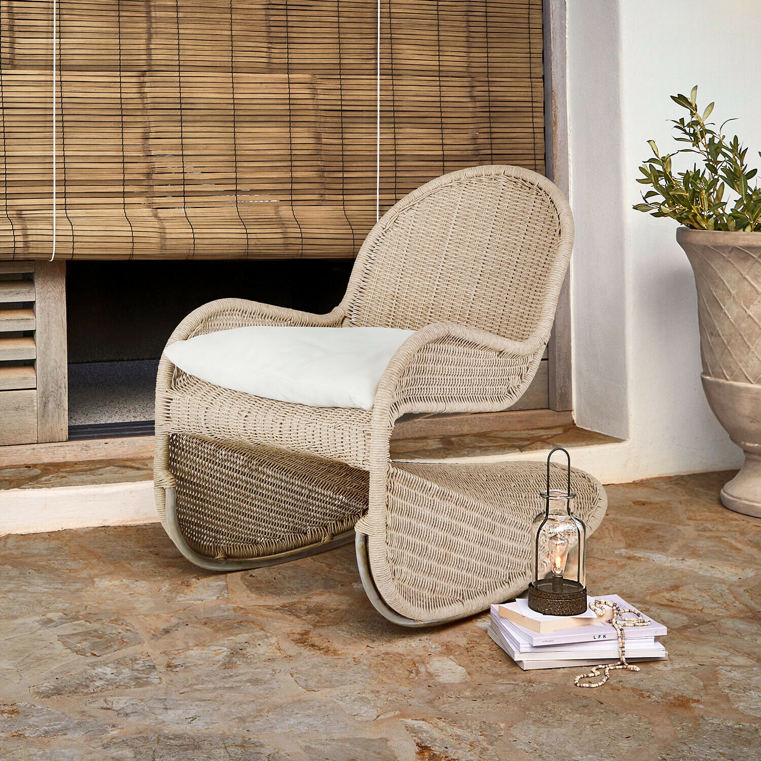 Fauteuil à bascule Fernleigha Fauteuil en rotin beige avec coussin blanc sur sol en pierre devant un store en bambou