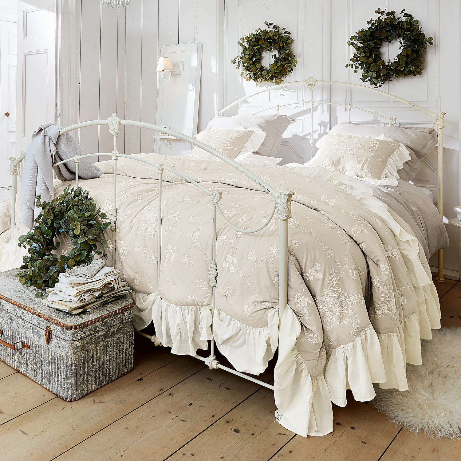 Courtepointe Clarissa Parure de lit beige brodée avec volants sur lit en métal blanc dans une chambre lumineuse décorée de couronnes vertes