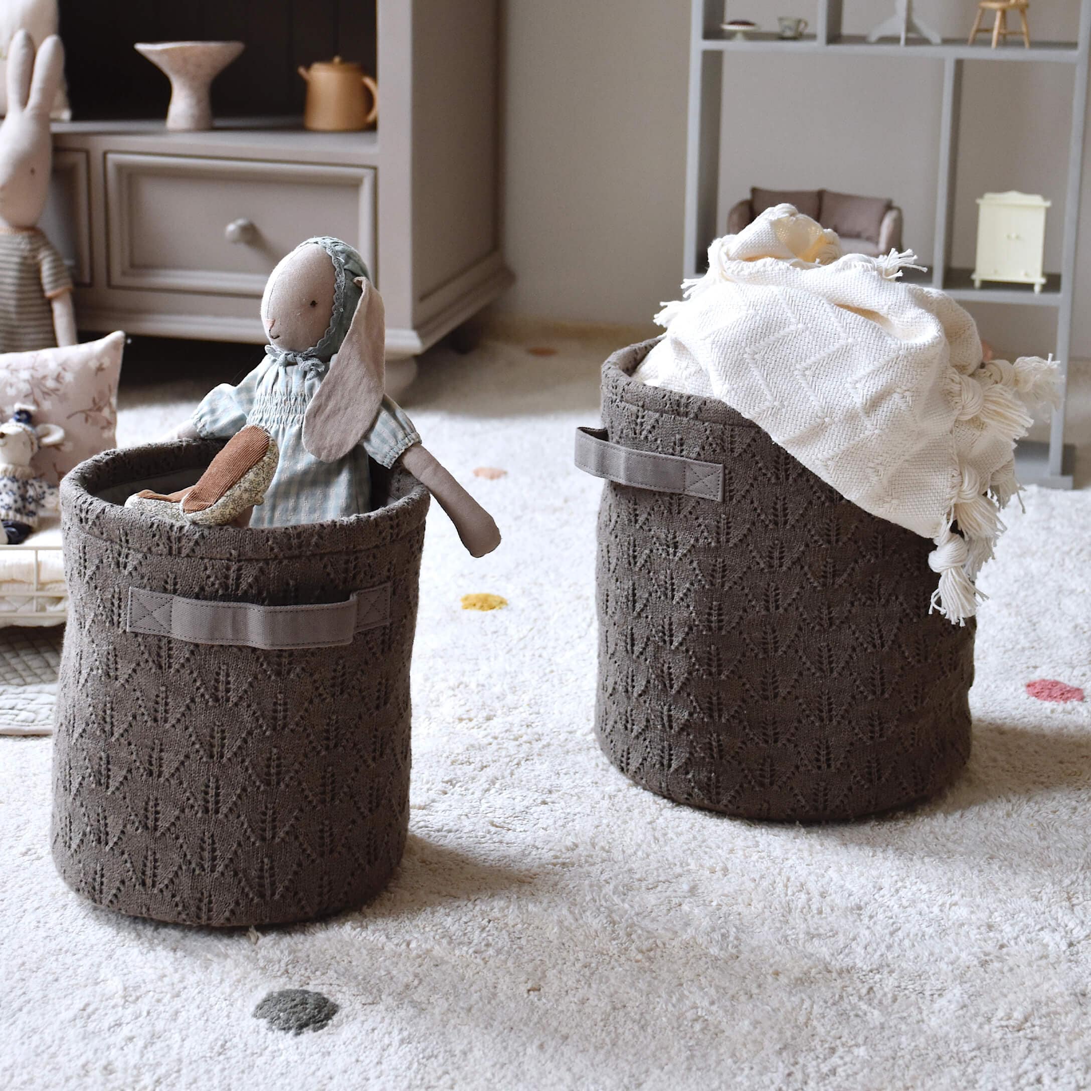 Paniers en tricot marron avec poignées, remplis de jouets et d'une couverture, posés sur un tapis clair