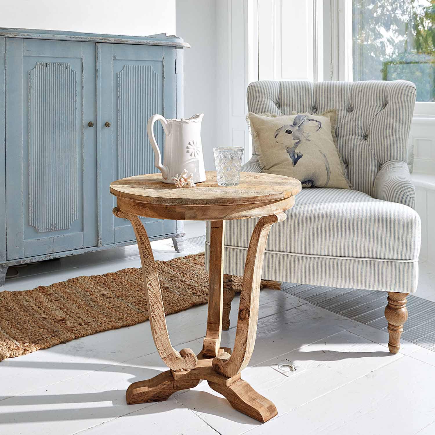 Table ronde en bois de manguier naturel dans un salon lumineux avec fauteuil rayé et buffet bleu clair