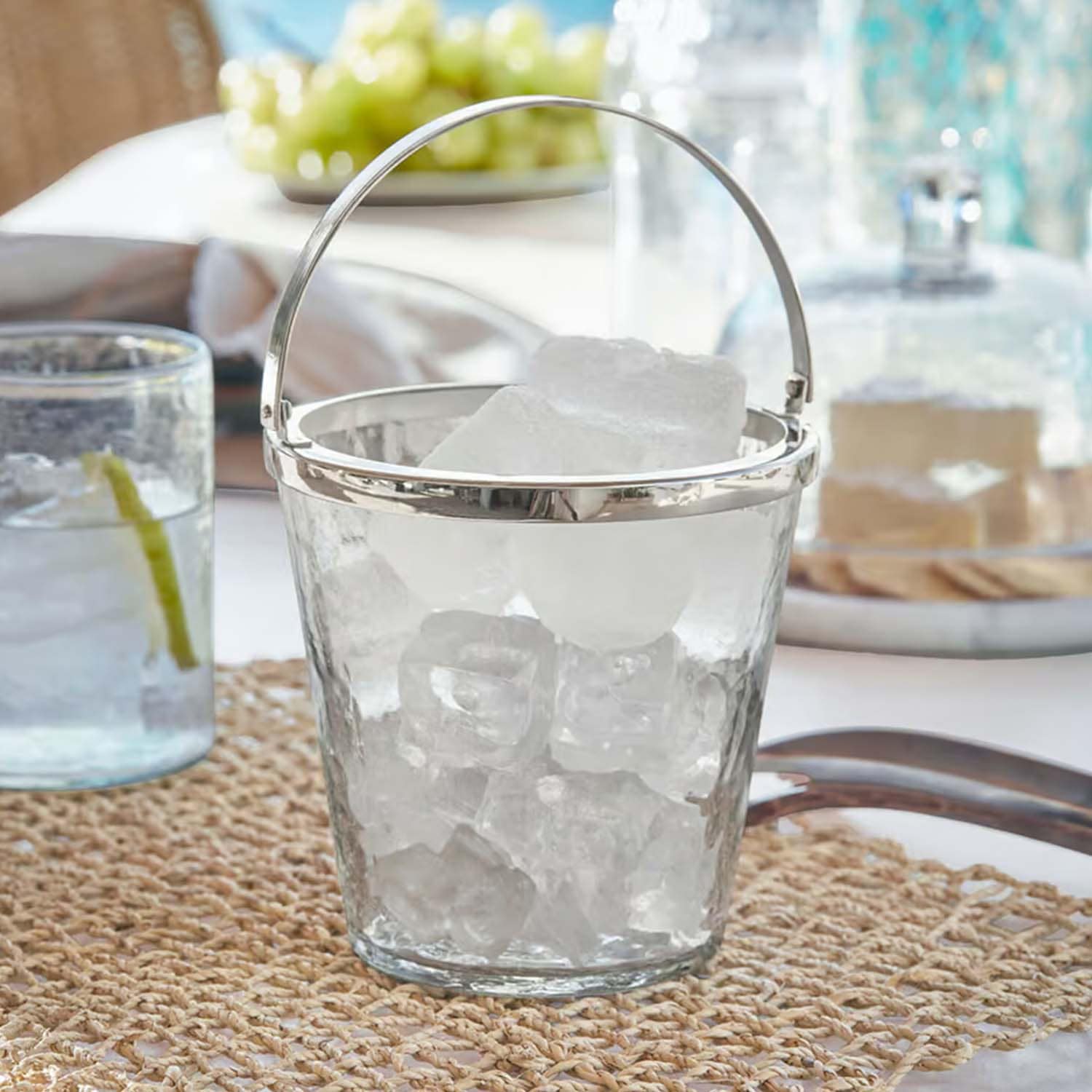 Seau à glaçons en verre transparent avec poignée en métal sur table avec napperon tressé