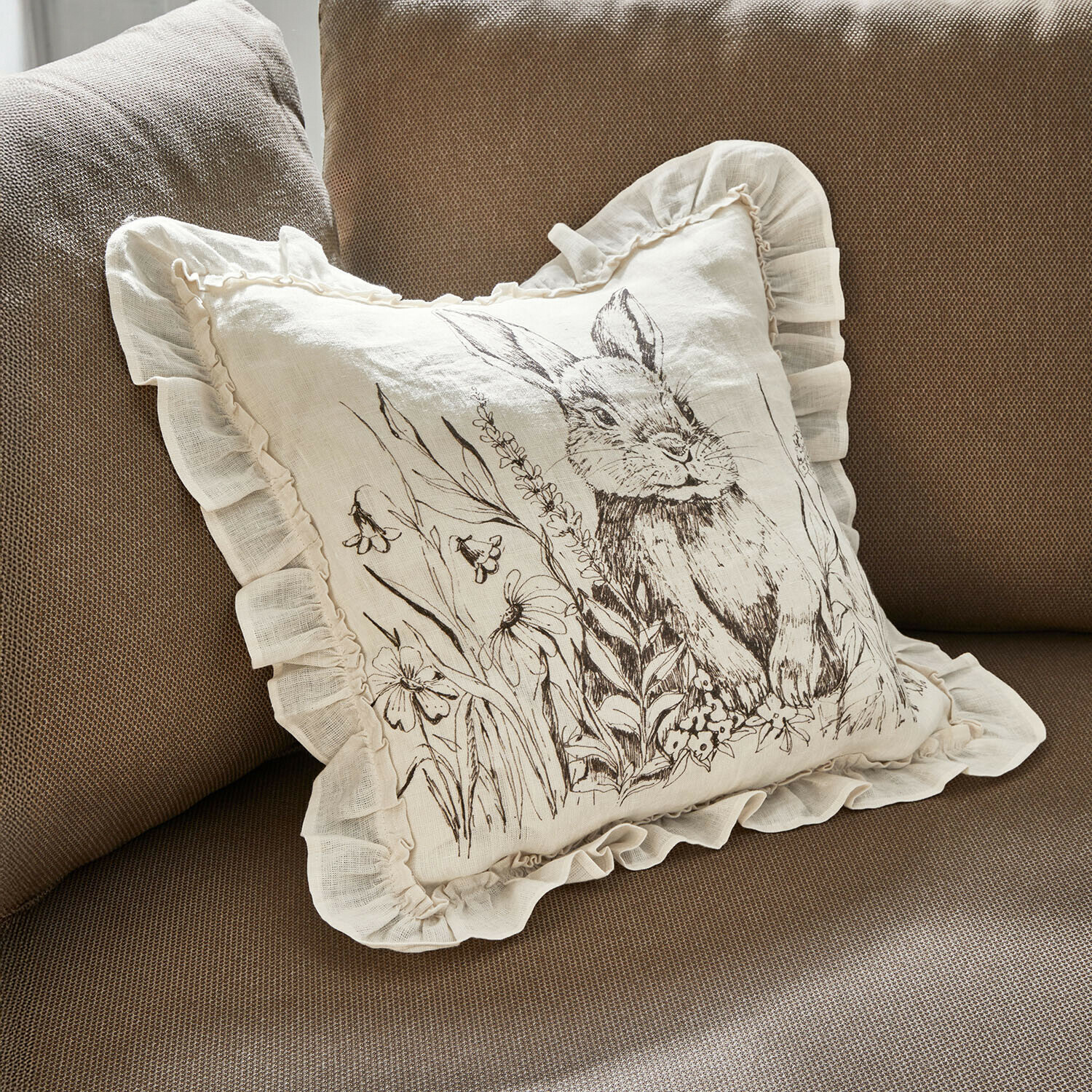Housse de coussin Brionelle Coussin en lin beige avec volant et motif lapin entouré de fleurs sur canapé marron