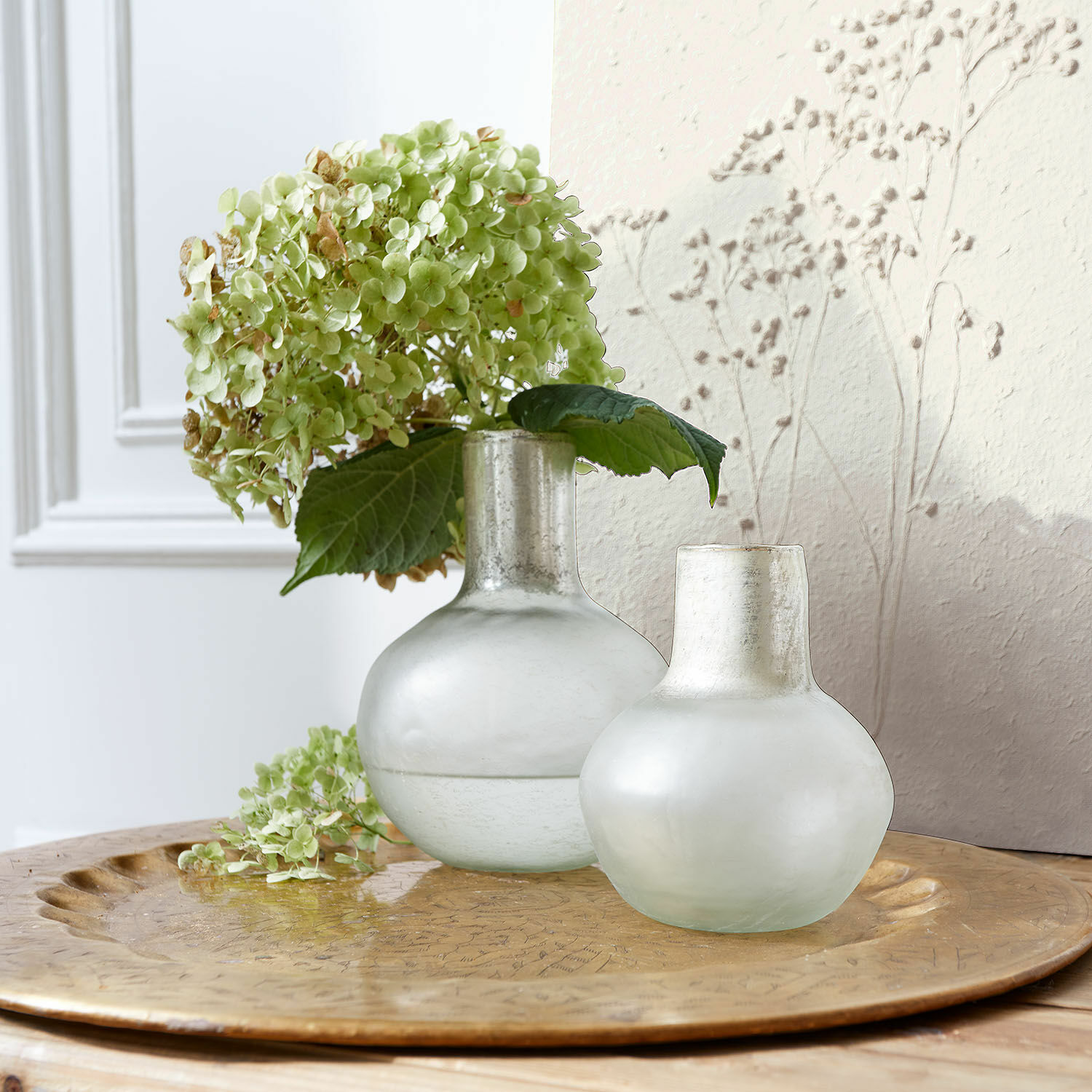 Lot de 2 vases Nazira Deux vases en verre givré couleur lait sur un plateau doré avec des fleurs vertes d'hortensia