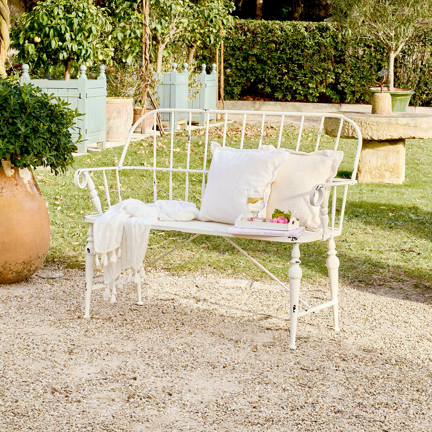 Banc de jardin en métal blanc avec coussins et plaid sur terrasse en gravier dans un jardin verdoyant