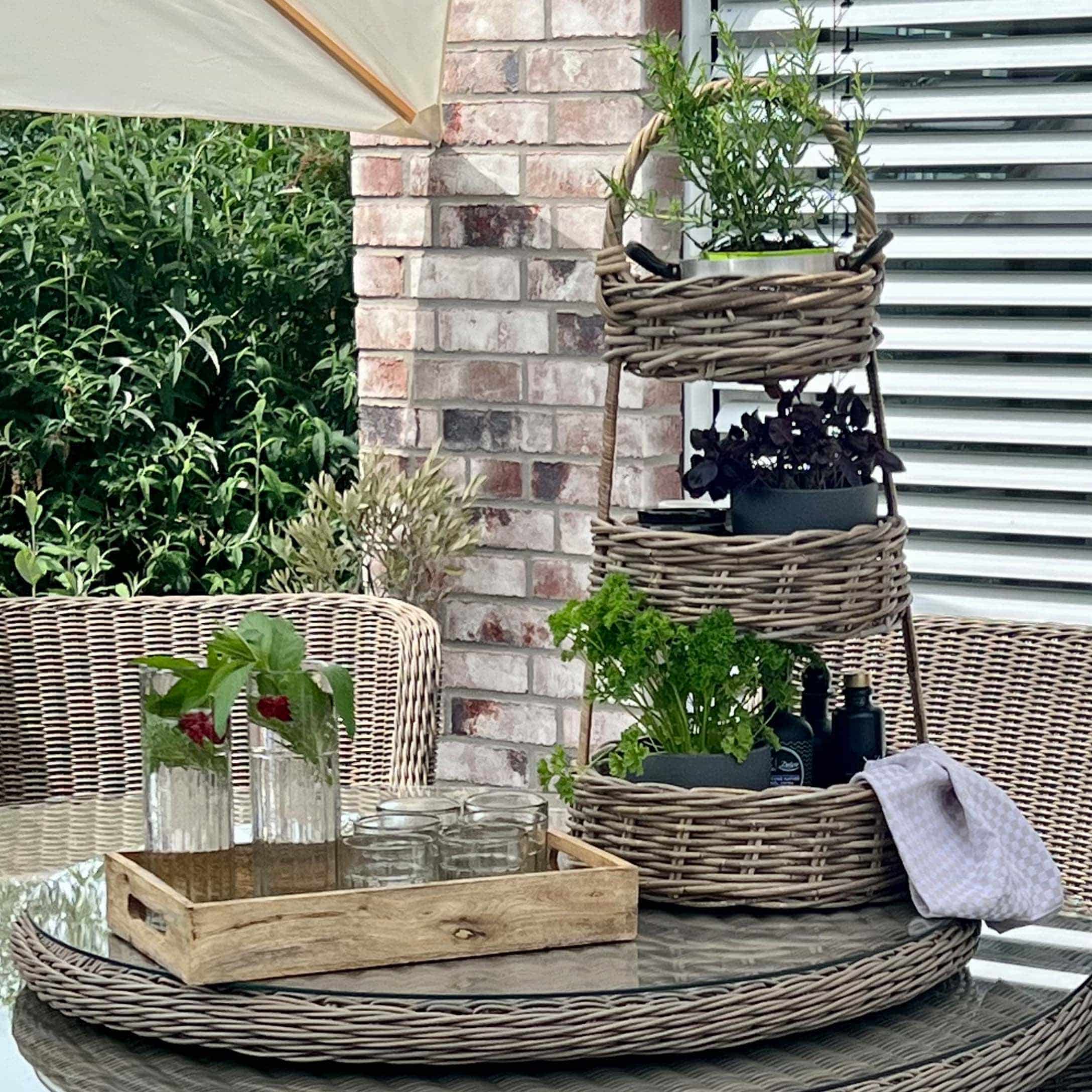 Étagère à trois niveaux en osier avec plantes vertes sur table en verre sur terrasse avec mobilier en rotin