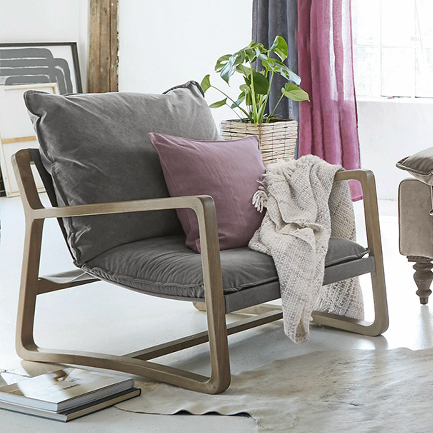 Fauteuil Jowna Fauteuil en tissu gris avec cadre en bois, coussin violet et plaid beige dans un salon lumineux