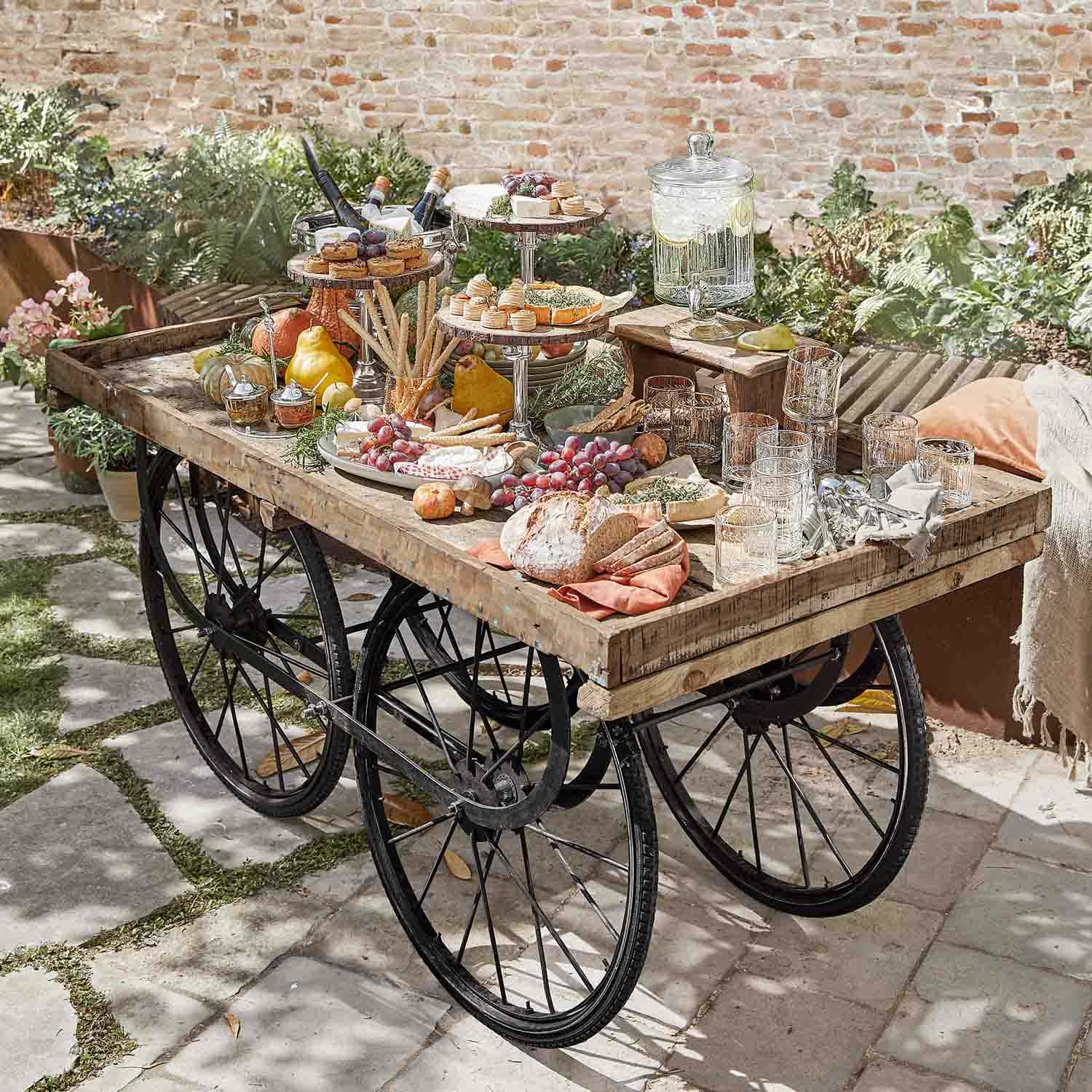 Chariot en bois rustique avec roues noires chargé de snacks et boissons dans un jardin ensoleillé