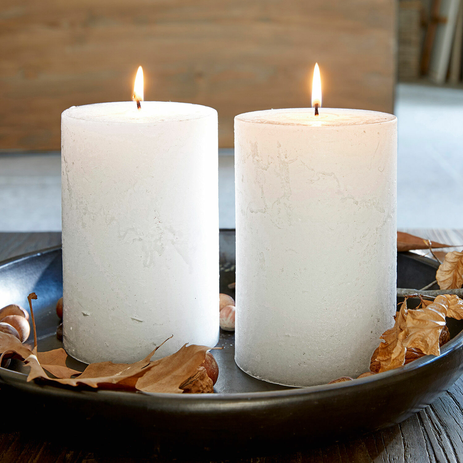 Lot de 2 bougies Oviedo Deux bougies blanches en cire sur un plateau noir avec feuilles sèches et noisettes, décoration automnale