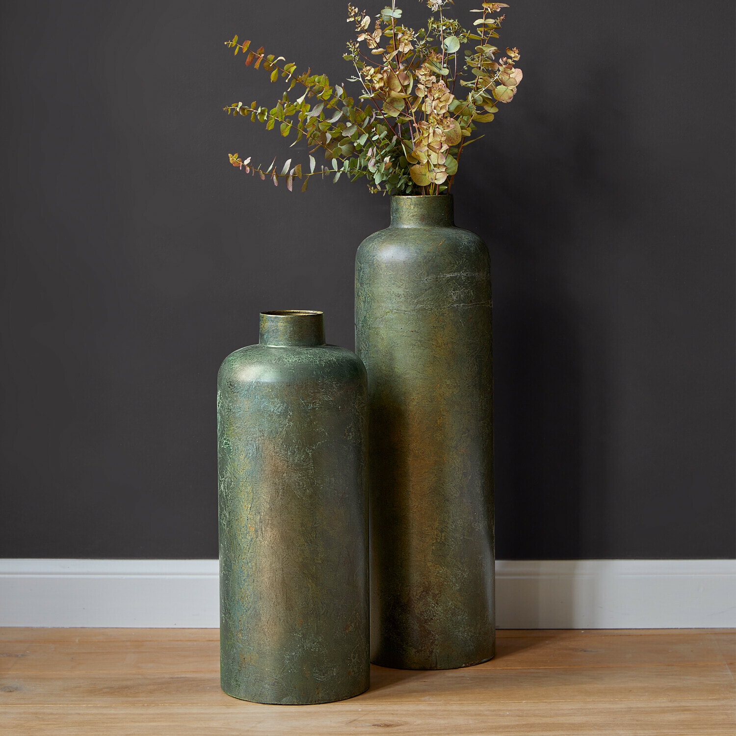 Lot de 2 vases Darwani Deux grands vases décoratifs verts en métal posés sur un sol en bois avec des feuilles séchées