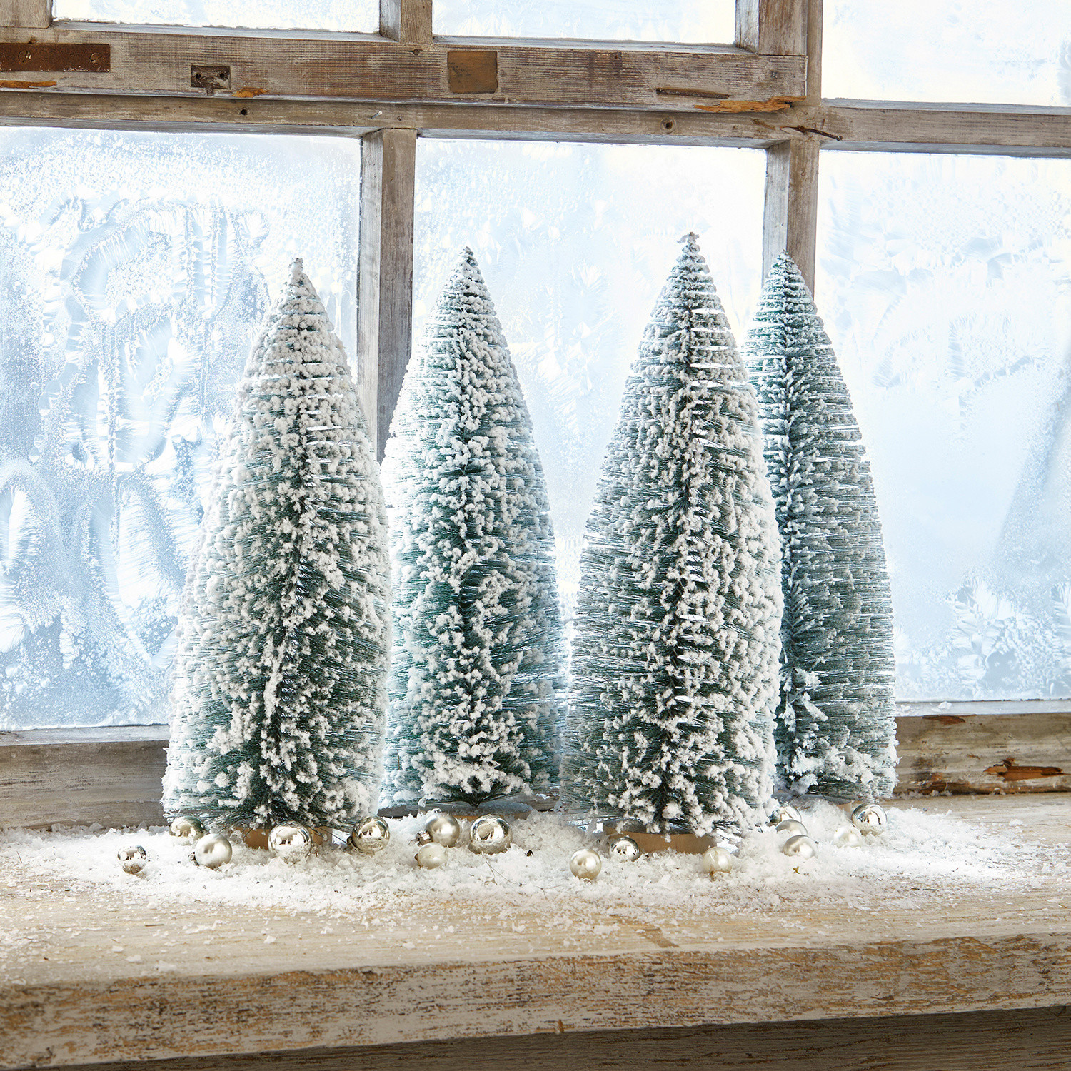 Lot de 4 arbres décoratifs Landscape Quatre sapins coniques verts en plastique avec neige blanche sur un rebord de fenêtre en bois décoré de boules argentées