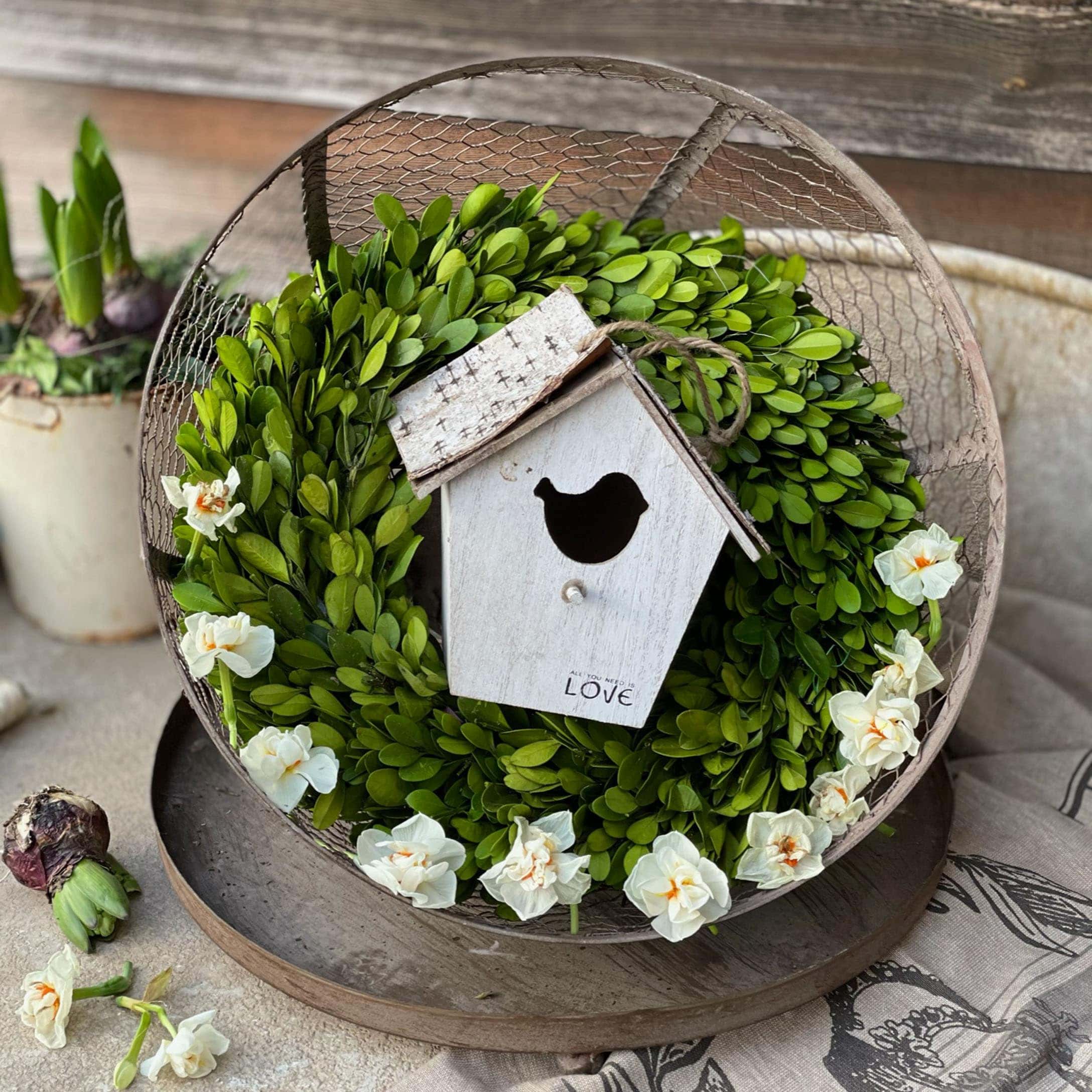Couronne décorative en feuilles vertes et fleurs blanches avec nichoir en bois sur une table rustique