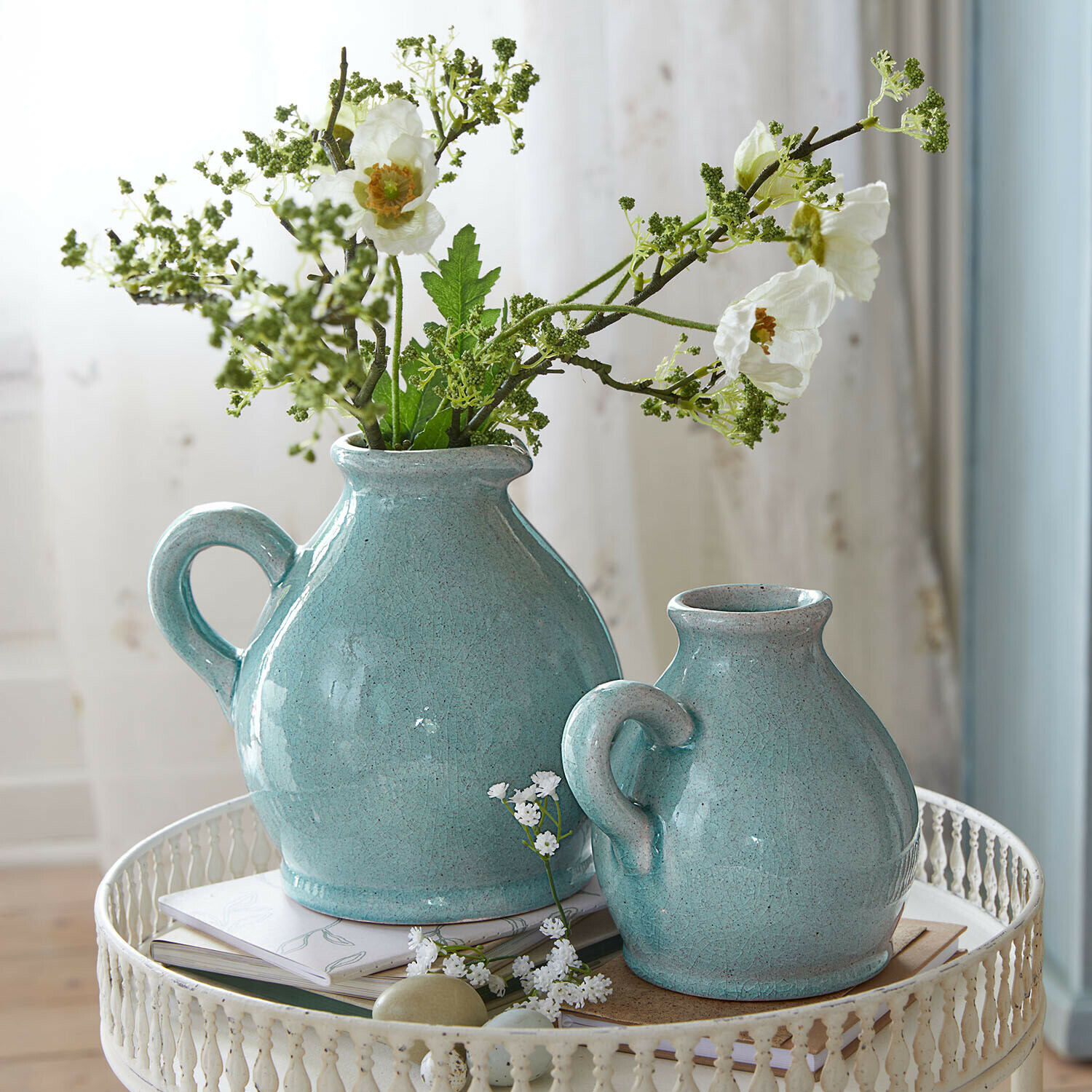 Deux pichets en céramique turquoise avec anses sur une table en métal blanc avec fleurs et livres