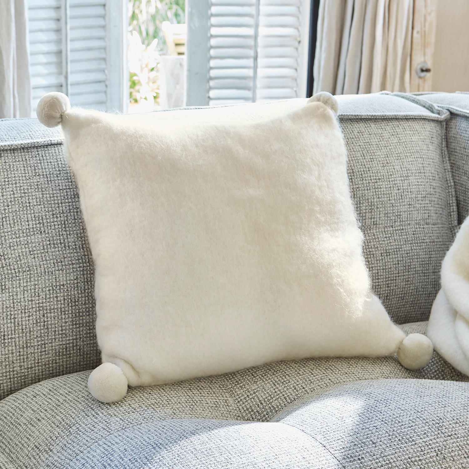 Coussin Mencia Coussin blanc doux en laine avec pompons sur canapé gris, décoration intérieure cosy