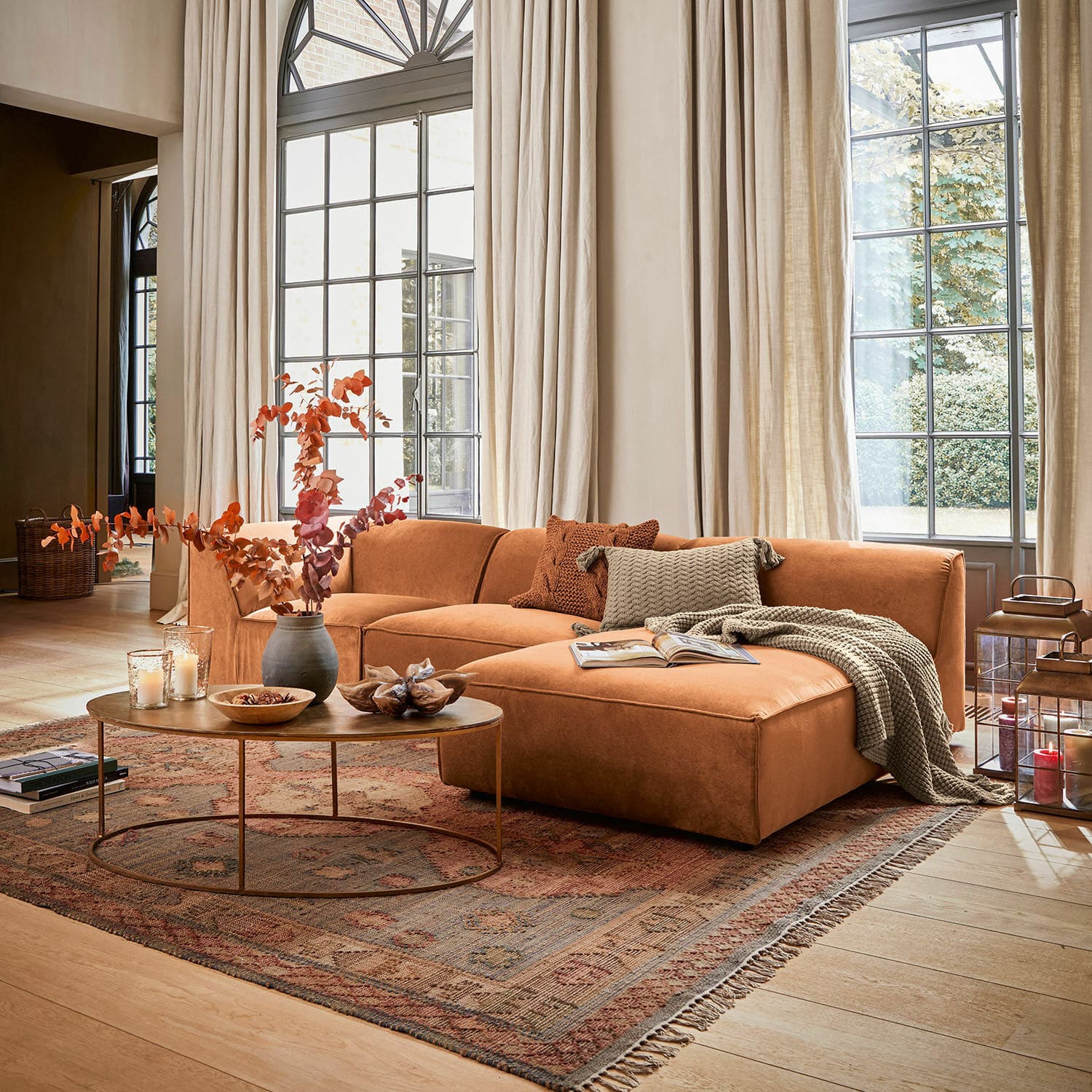 Canapé McLean Canapé d'angle couleur brique avec coussins et plaid dans un salon lumineux aux grandes fenêtres