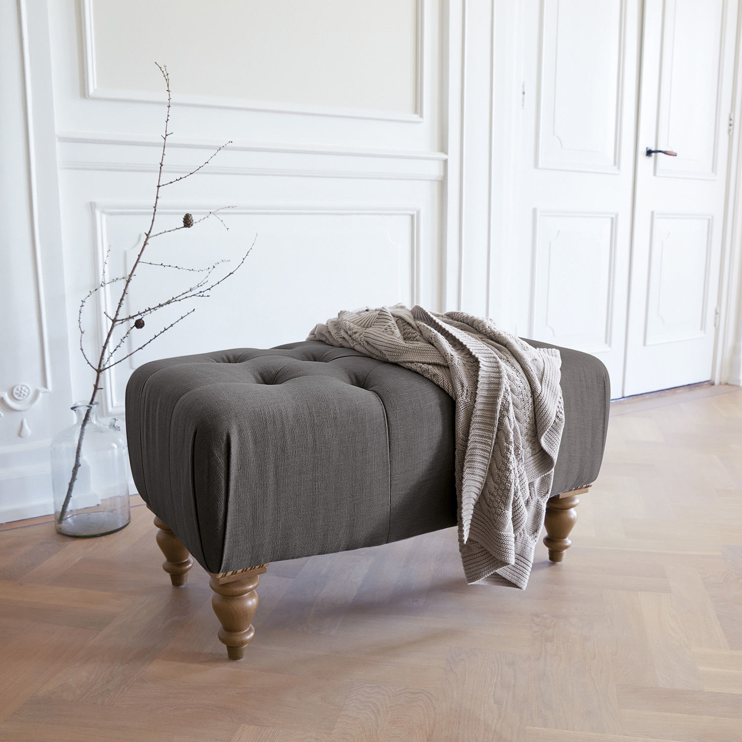 Tabouret Durance Pouf rembourré gris avec pieds en bois et couverture beige dans un intérieur lumineux