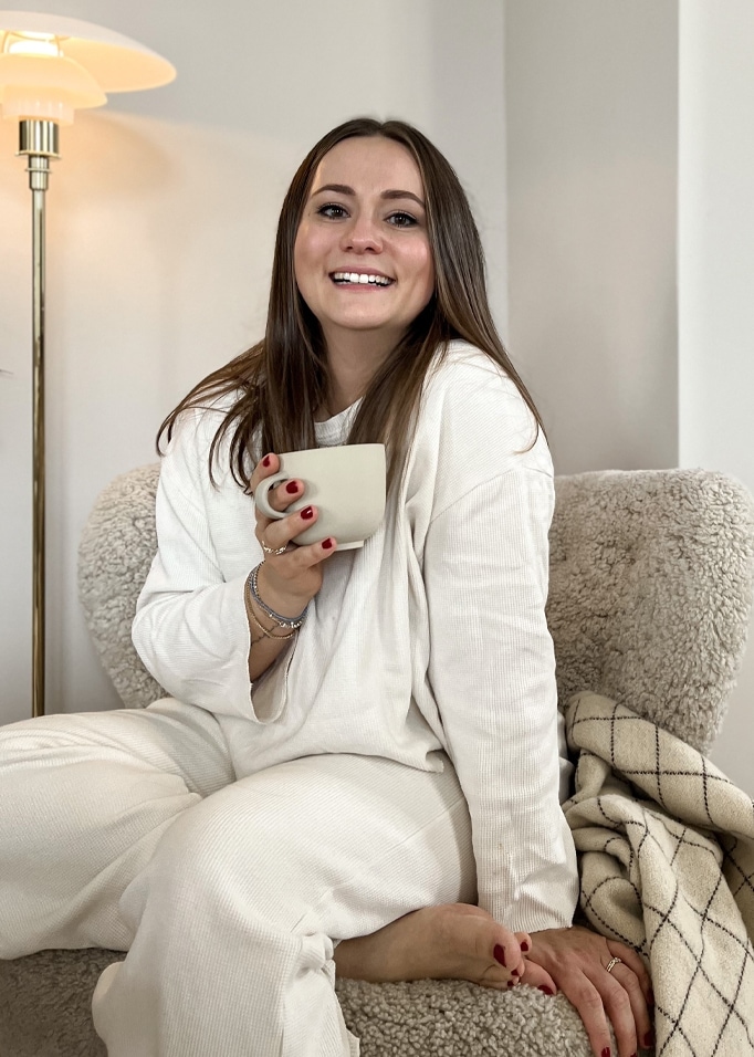 Femme en tenue blanche confortable assise sur un fauteuil avec une tasse, ambiance chaleureuse à la maison