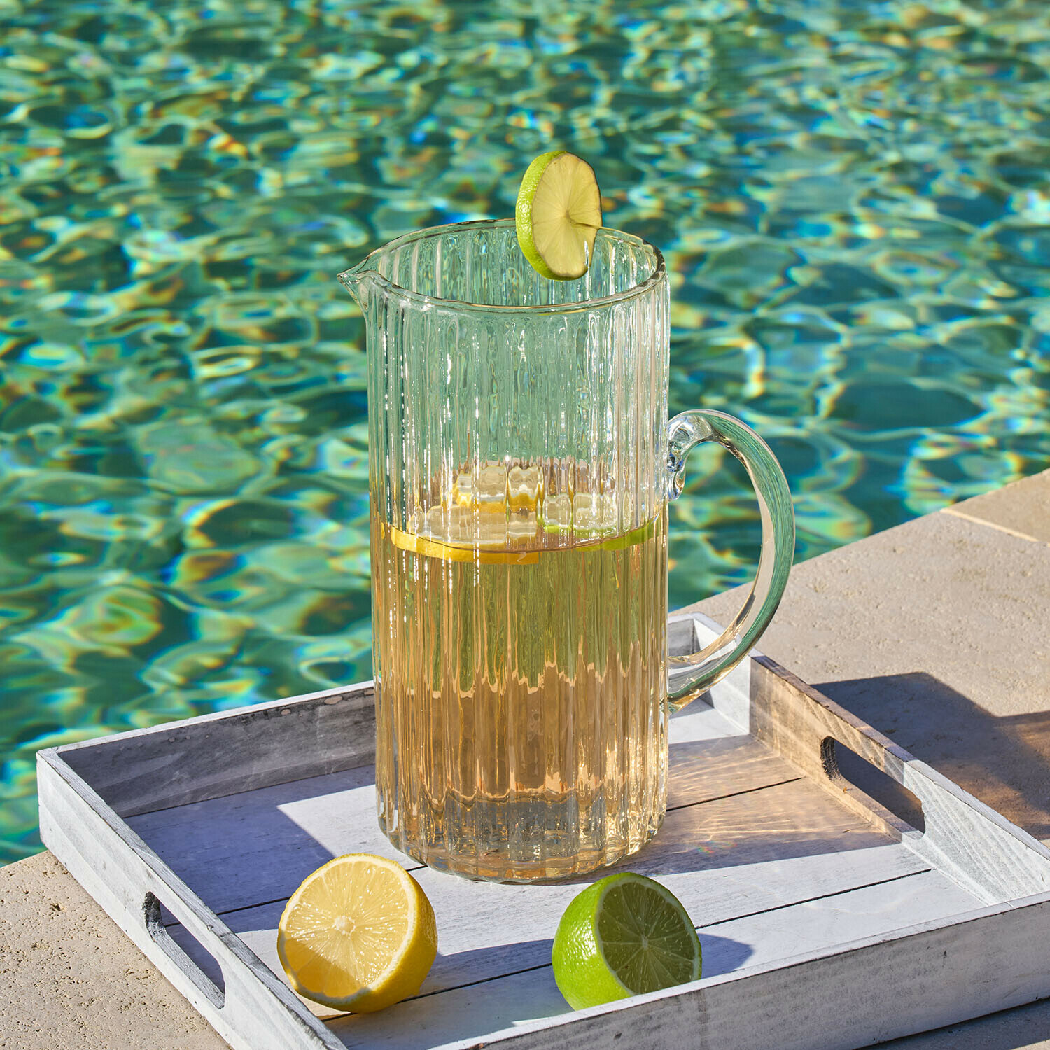 Carafe en verre transparent avec boisson et tranche de citron vert sur plateau blanc au bord de la piscine