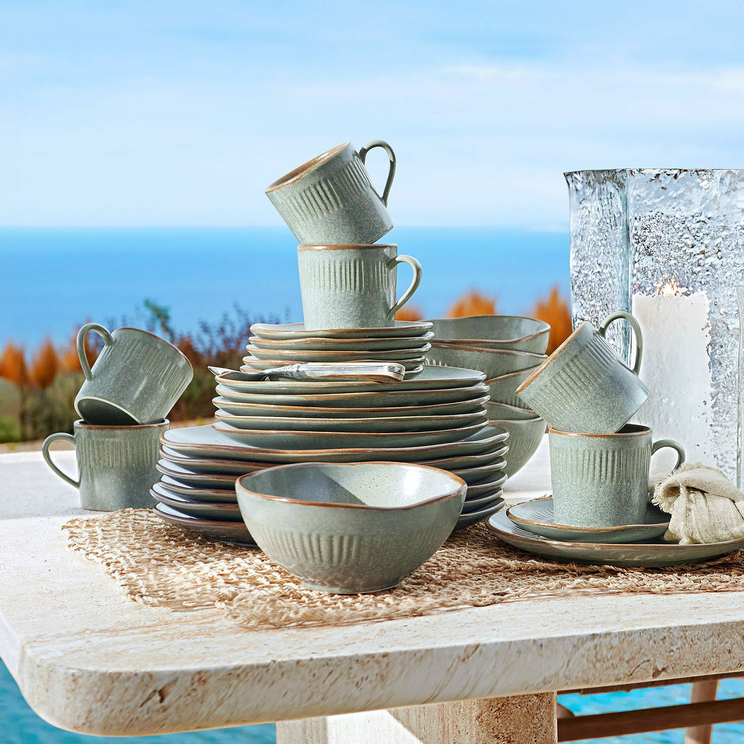 Service 30 pièces Périers Service de table en céramique grise avec tasses et assiettes empilées sur une table extérieure face à la mer