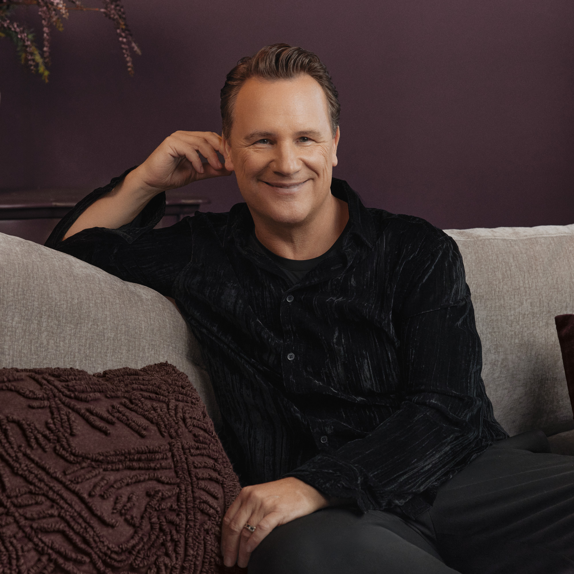 Homme souriant en chemise noire en velours assis sur un canapé gris clair avec une couverture bordeaux