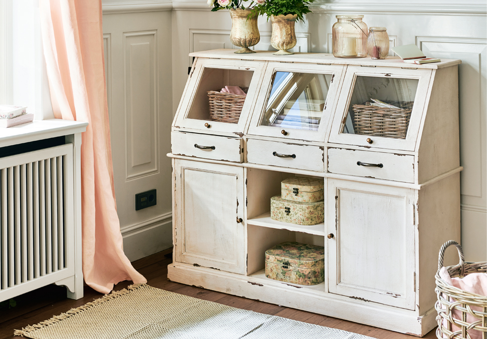 Buffet en bois blanc style shabby chic avec tiroirs vitrés et décorations dans une pièce lumineuse.