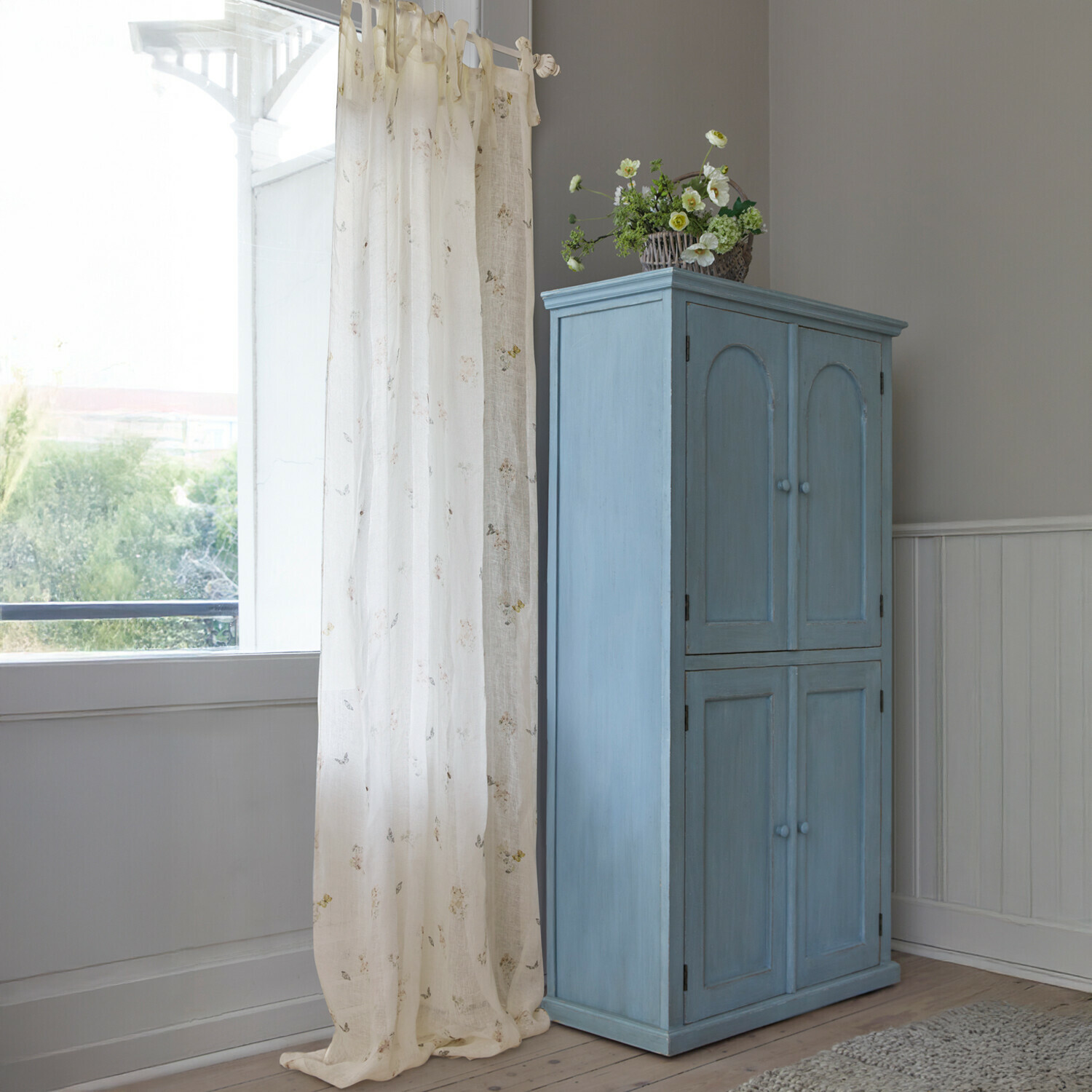 Rideau Salénaïs Armoire en bois bleue à quatre portes avec panier de fleurs décoratif dans une pièce lumineuse