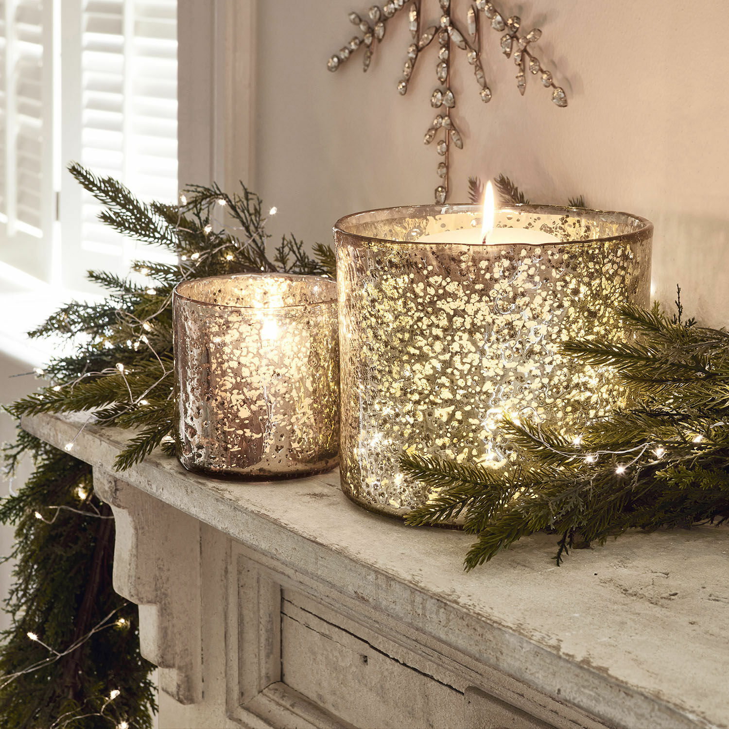 Lot de 2 photophores Samuel Deux photophores en verre effet argent vieilli sur une cheminée avec branches vertes et guirlandes lumineuses