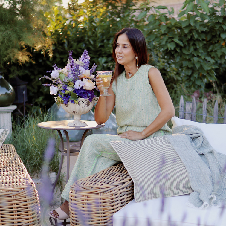 Femme en robe verte assise sur un meuble en osier dans un jardin avec bouquet de fleurs et verre de vin