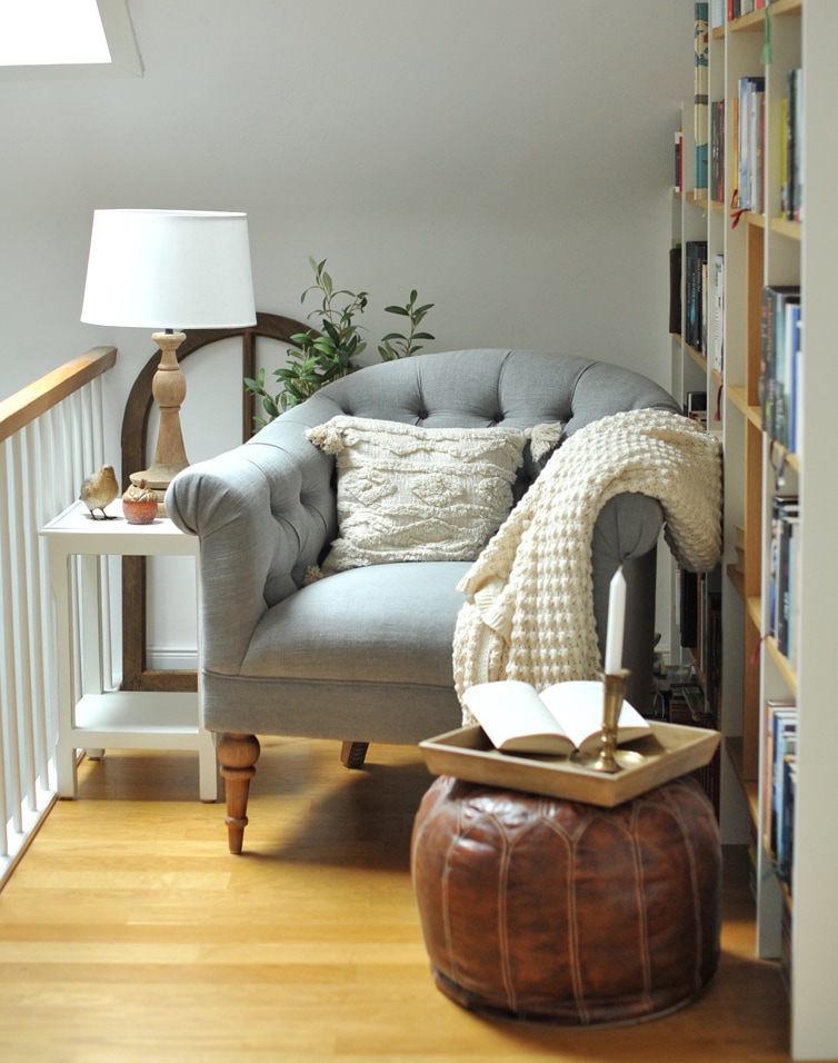 Fauteuil gris en tissu avec coussin et plaid crème, pouf en cuir marron et lampe sur table blanche